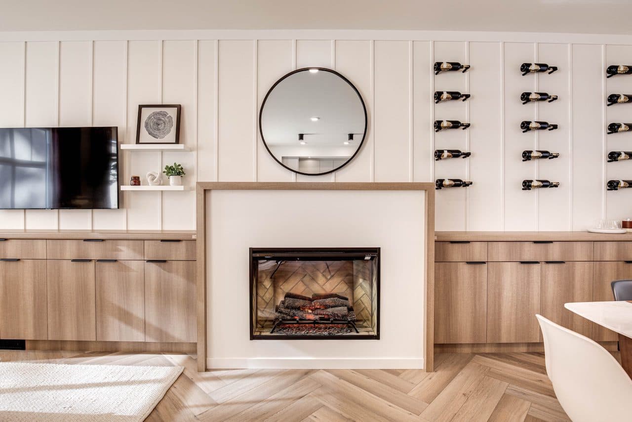 The Loop – Living and dining feature wall with a fireplace, round mirror, mounted TV, wood cabinetry, and a wall-mounted wine bottle display.