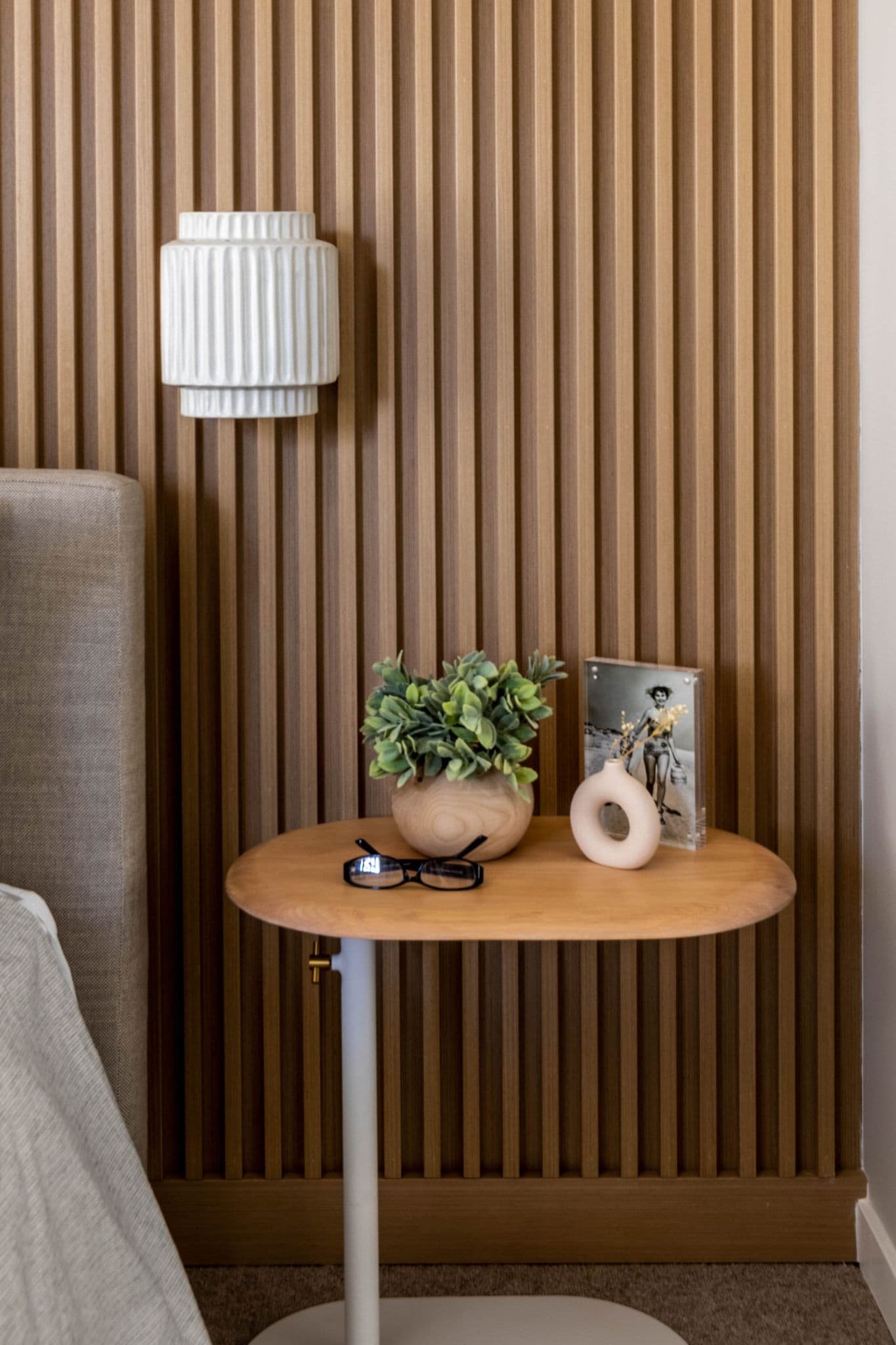 The Loop – Bedside vignette with a wood-slat accent wall, wall sconce, small oval side table holding a plant, glasses, and a framed photo.