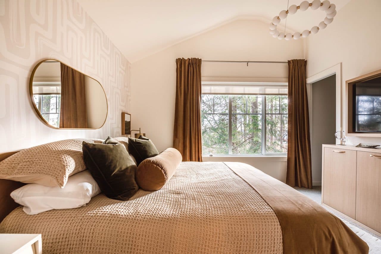 The Loop – Bedroom with an oval mirror, patterned wallpaper, tan bedding with olive velvet pillows, and a window looking out to trees.