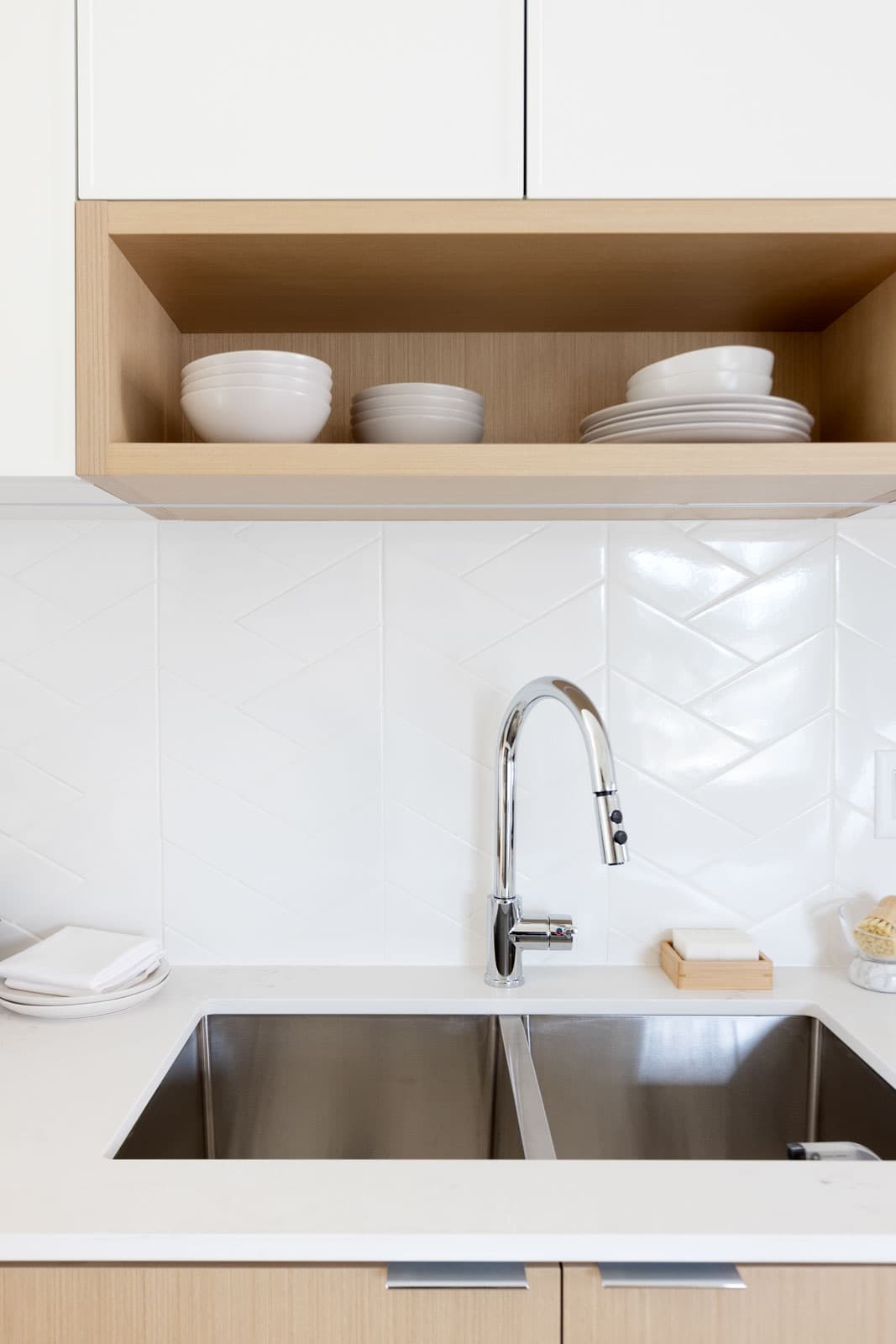 The Loop – Close-up of a kitchen sink and faucet with a herringbone tile backsplash and an open wood shelf holding stacked white dishware above.