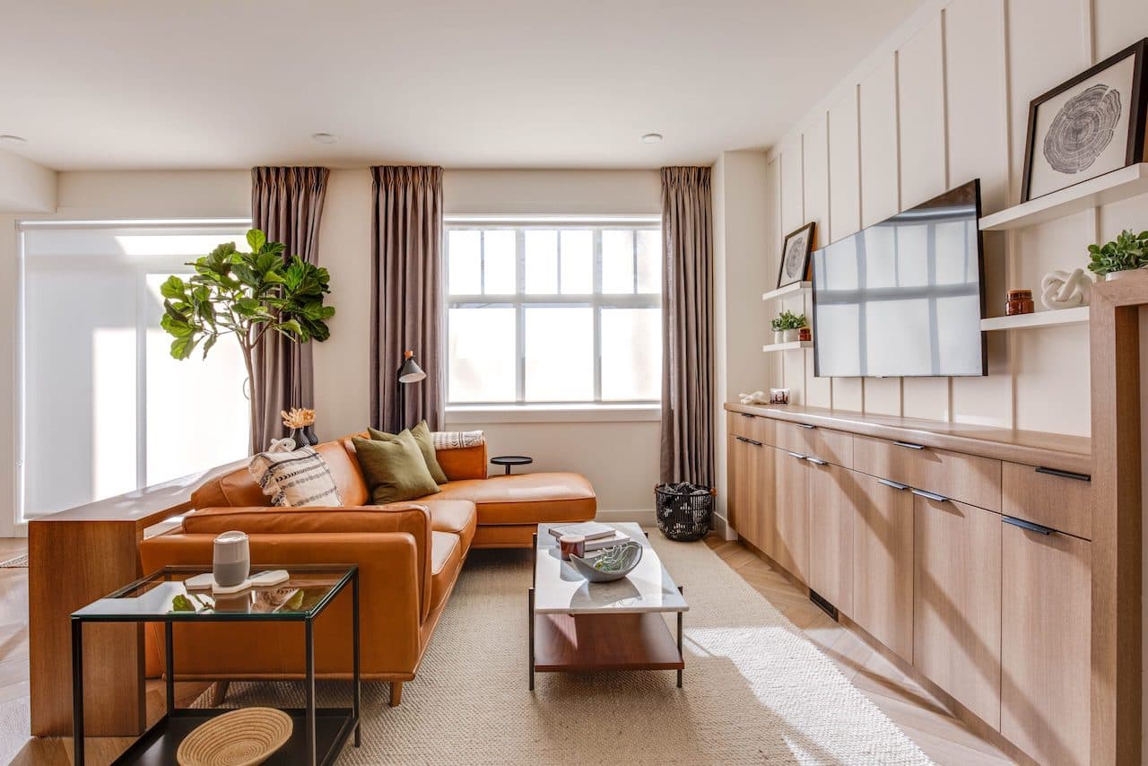 The Loop – Living room with a cognac leather sectional sofa, glass coffee table, built-in wood cabinetry, and a wall-mounted TV.