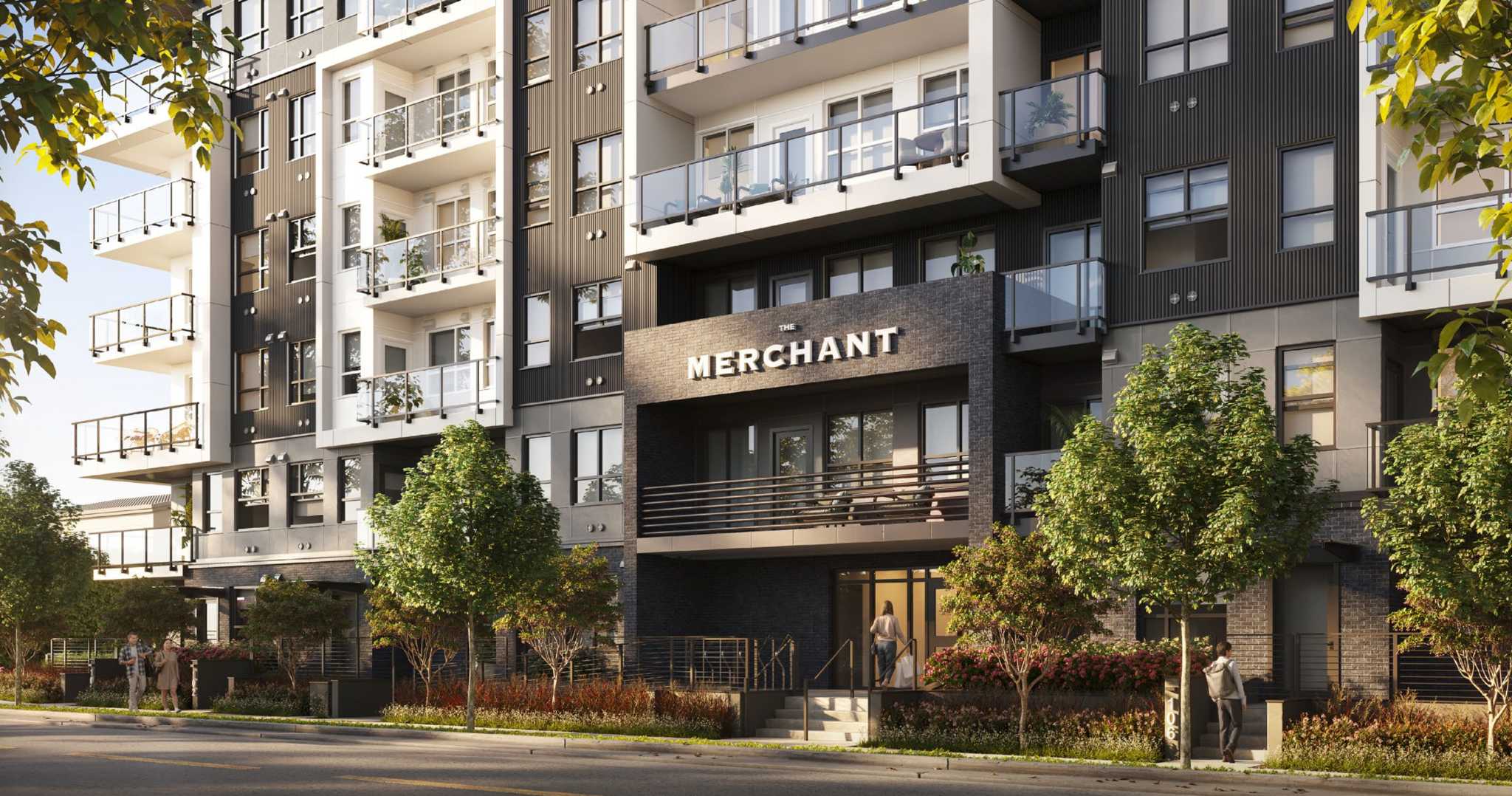 The Merchant – Street-level view of the building entrance with a dark brick facade, THE MERCHANT signage, balconies above, and landscaped planters along the sidewalk.