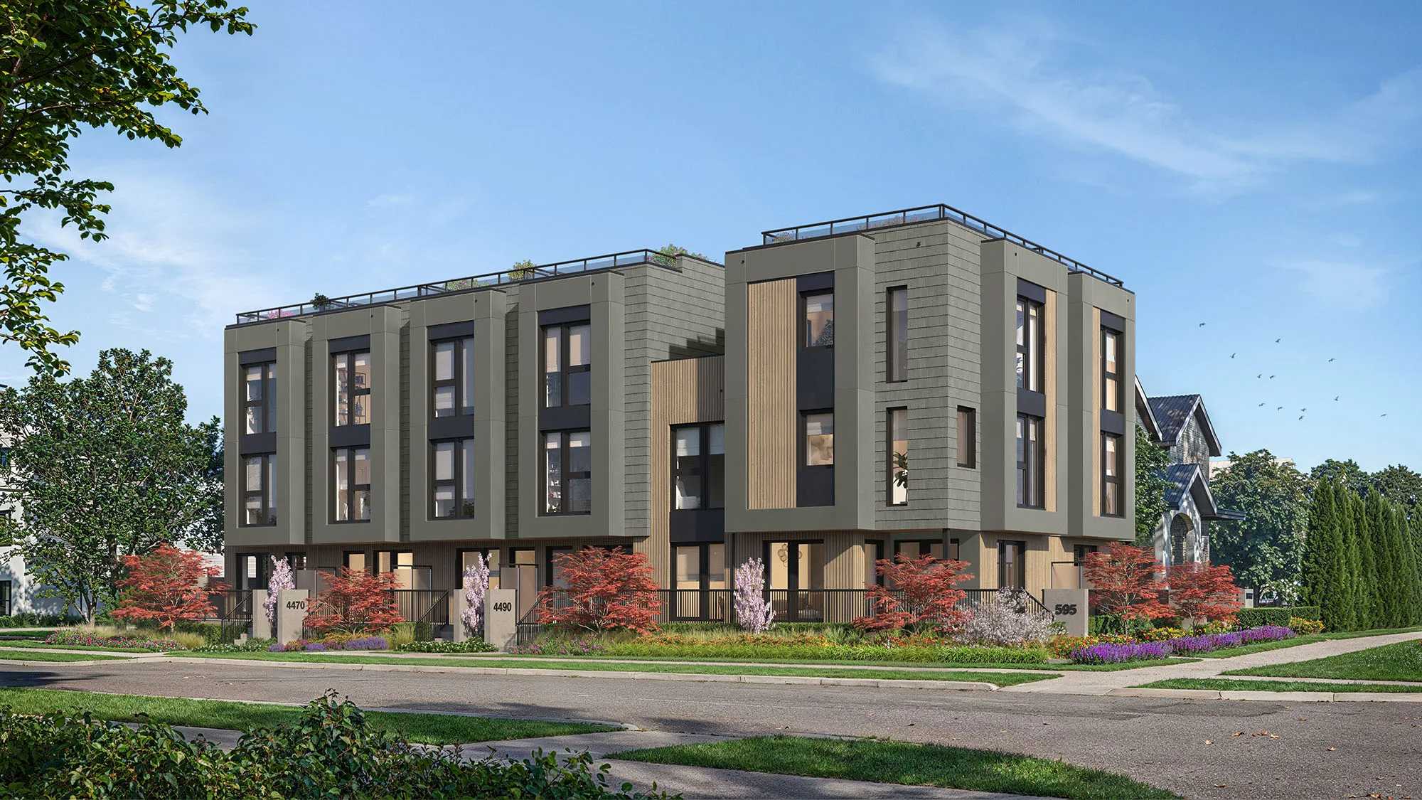 Nolia – A modern three-story multi-family townhouse complex featuring grey horizontal siding, light wood vertical accents, and private rooftop terraces. The building sits along a paved suburban street bordered by lush landscaping, blooming purple flowers, and red foliage under a bright daytime sky.