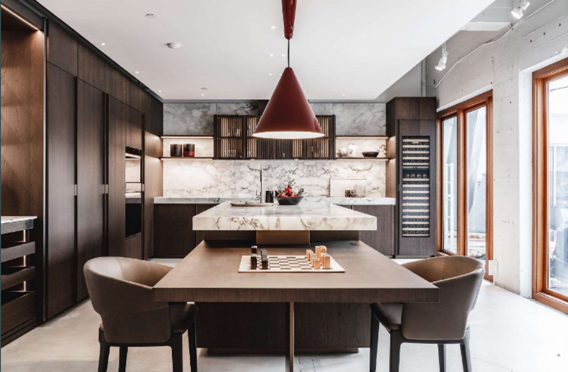 Ashlyn – A high-end modern kitchen featuring dark wood cabinetry, an integrated tall wine fridge, and striking white marble backsplashes and countertops. In the foreground, an attached wood dining table is set with a chessboard, accompanied by leather-upholstered chairs beneath a large conical pendant light.