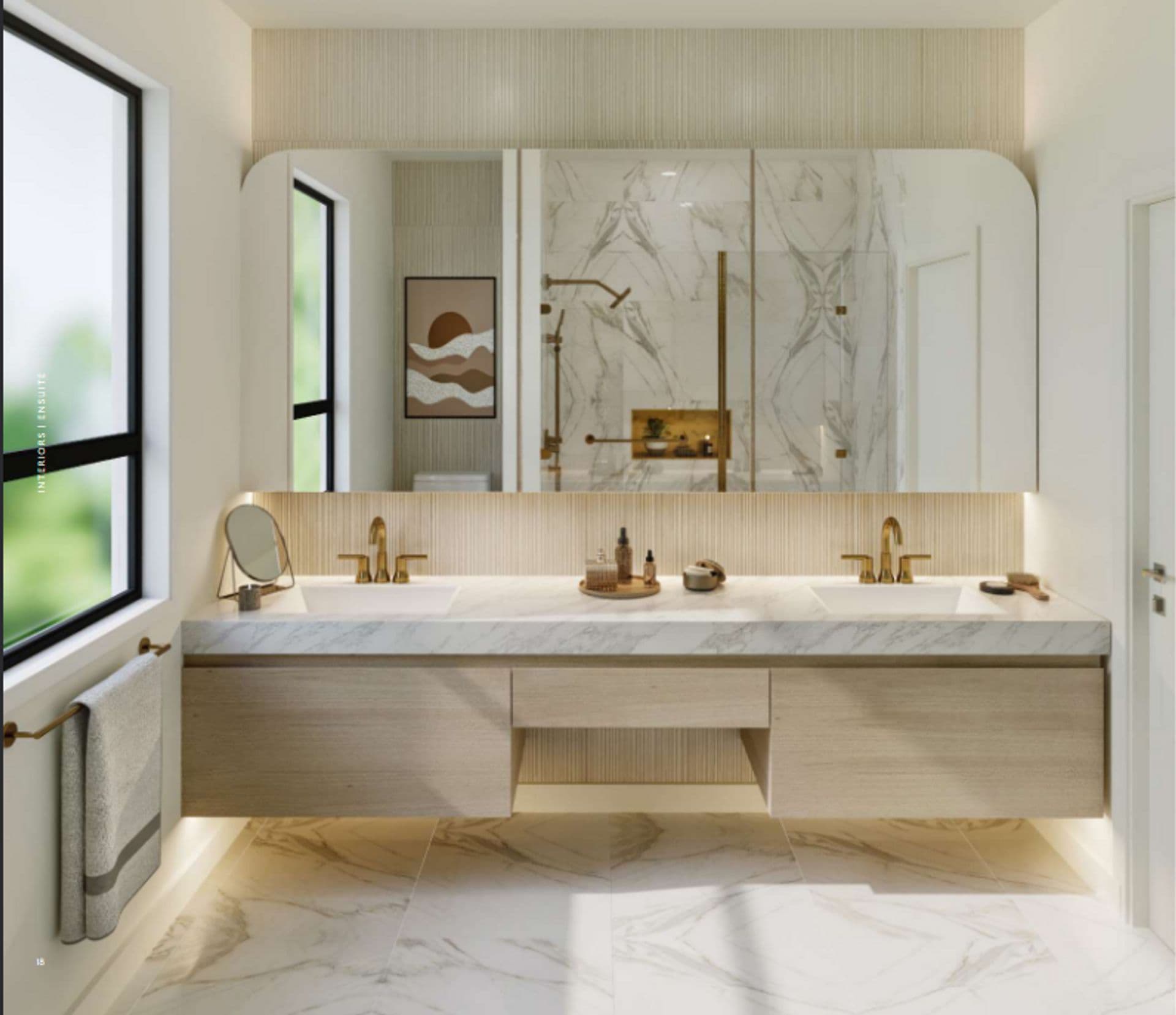 Ashlyn – A bright, modern ensuite bathroom featuring a floating light-wood double vanity with marble countertops and brushed gold fixtures. Large wide mirrors with warm integrated backlighting reflect a marble-tiled walk-in shower, while natural daylight streams in from a window onto matching marble floor tiles.