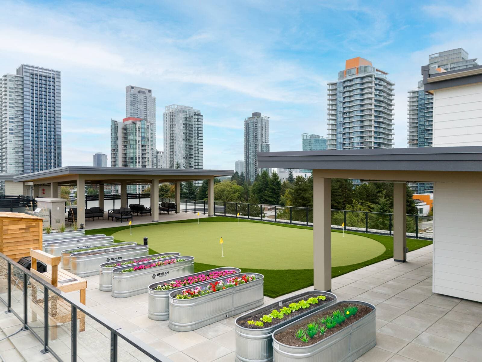Pura – A rooftop amenity terrace featuring a green putting green with yellow flags alongside galvanized metal planter boxes filled with flowers and greenery. Covered outdoor lounge seating areas frame the space, offering panoramic views of surrounding high-rise city towers under a bright daytime sky.
