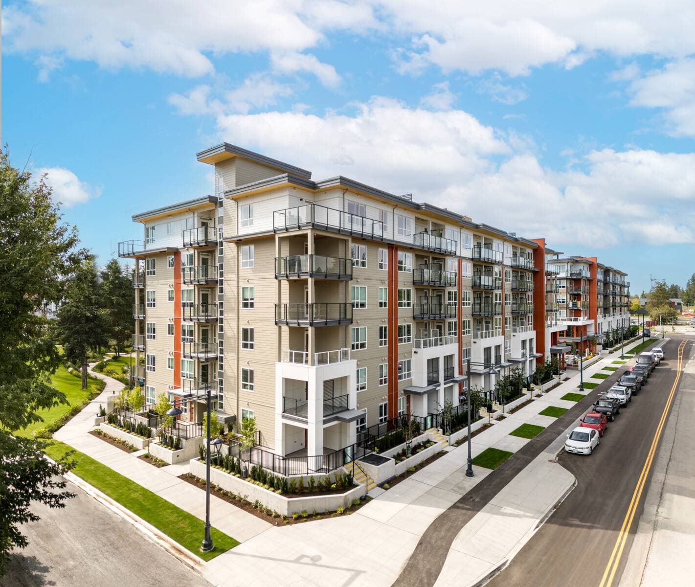 Pura – A modern multi-story residential building featuring light tan lap siding, warm orange vertical accents, and glass-railed balconies against a bright sky with fluffy clouds. Clean concrete sidewalks, street lamps, landscaped garden beds, and parallel-parked cars line the street below.
