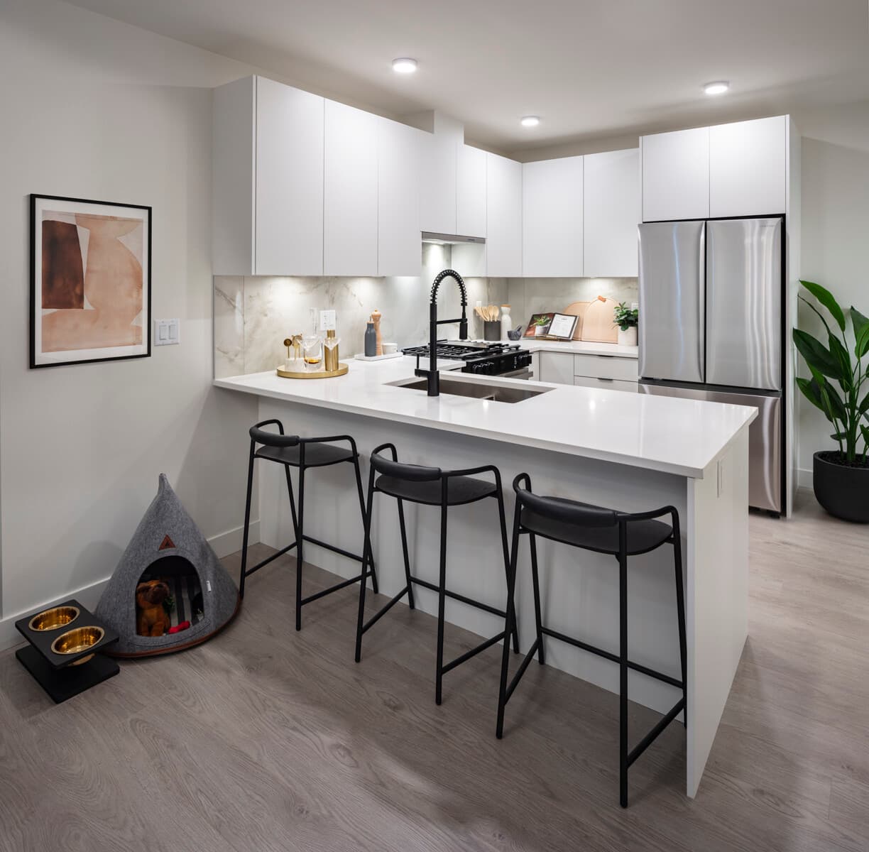 Pura – A modern, bright condo kitchen featuring white minimalist cabinetry, stainless steel appliances, and a marble-patterned backsplash. A large waterfall island with white quartz countertops is paired with three sleek black barstools, set against light wood flooring and staged with cozy pet accessories nearby.