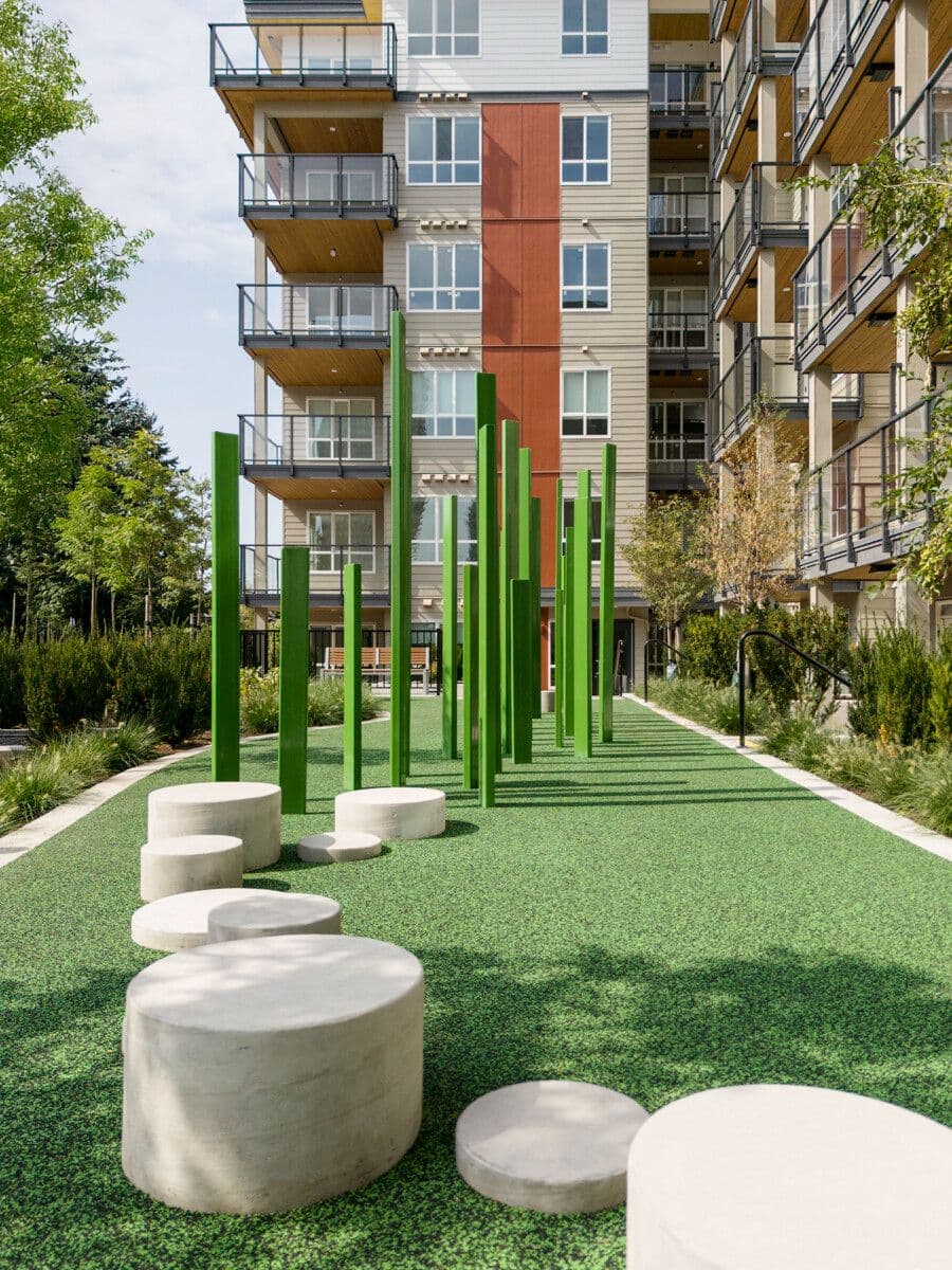 Pura – An outdoor outdoor play and leisure space features vibrant green vertical posts and circular concrete stepping stones set on green safety turf adjacent to a mid-rise residential building. The modern multi-story apartment complex stands in the background under bright daylight, surrounded by landscaped shrubs and trees.