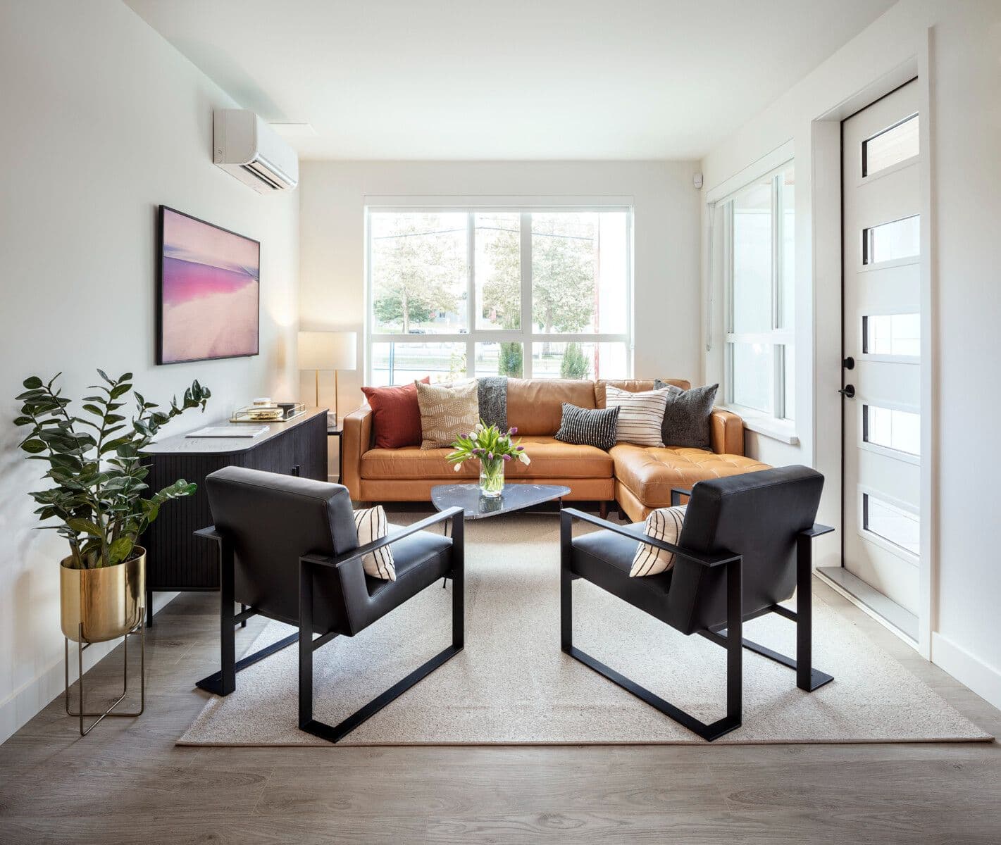 Pura – A bright, modern living room featuring a Cognac leather sectional sofa set against a large window, paired with two black armchairs and a dark wood credenza. Sunlight streams into the white-walled space, which is accented by a brass planter, framed artwork, and light wood laminate flooring.