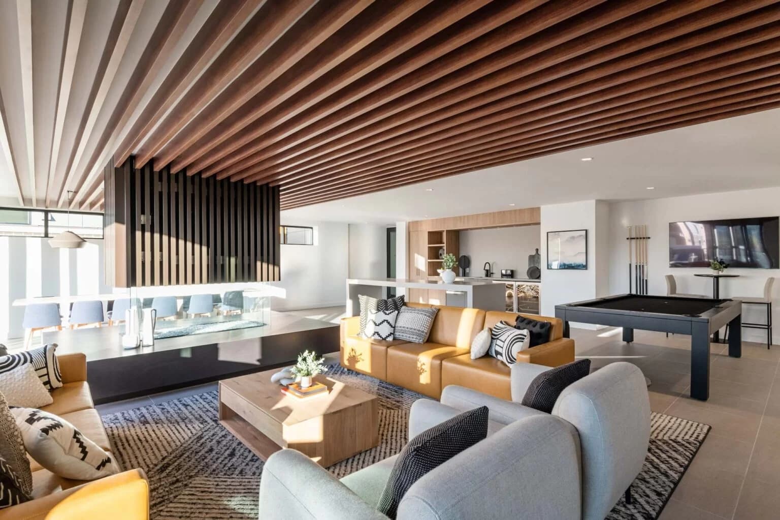 50 Electronic – A modern residential lounge features a central double-sided fireplace, rich wood slat ceiling detailing, and comfortable leather and fabric seating areas. In the background, the open space extends to a pool table, wet bar, and dining area bathed in natural sunlight.