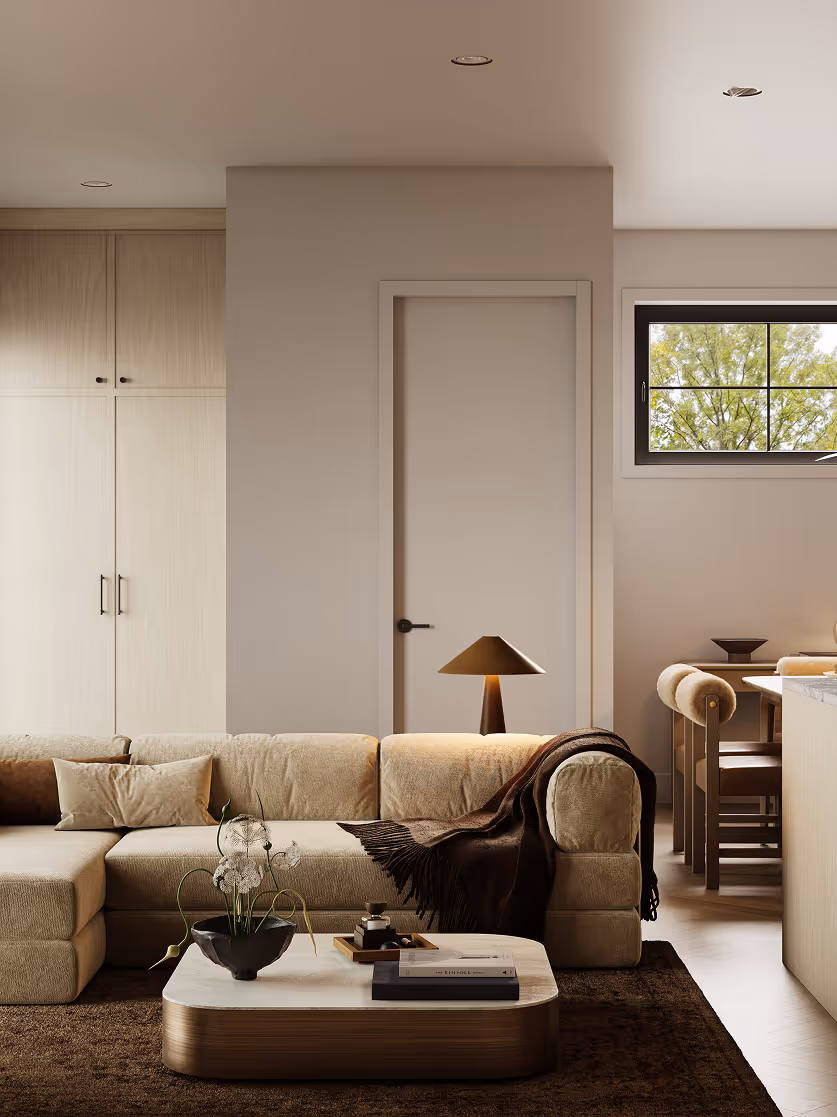 Wallace – A contemporary living area featuring a neutral-toned sectional sofa adorned with cushions and a brown throw blanket, set around a low coffee table with decorative accents. In the background, light wood built-in cabinetry, a dining space with chairs, and a window offering a view of lush green trees complement the soft, minimalist interior design.