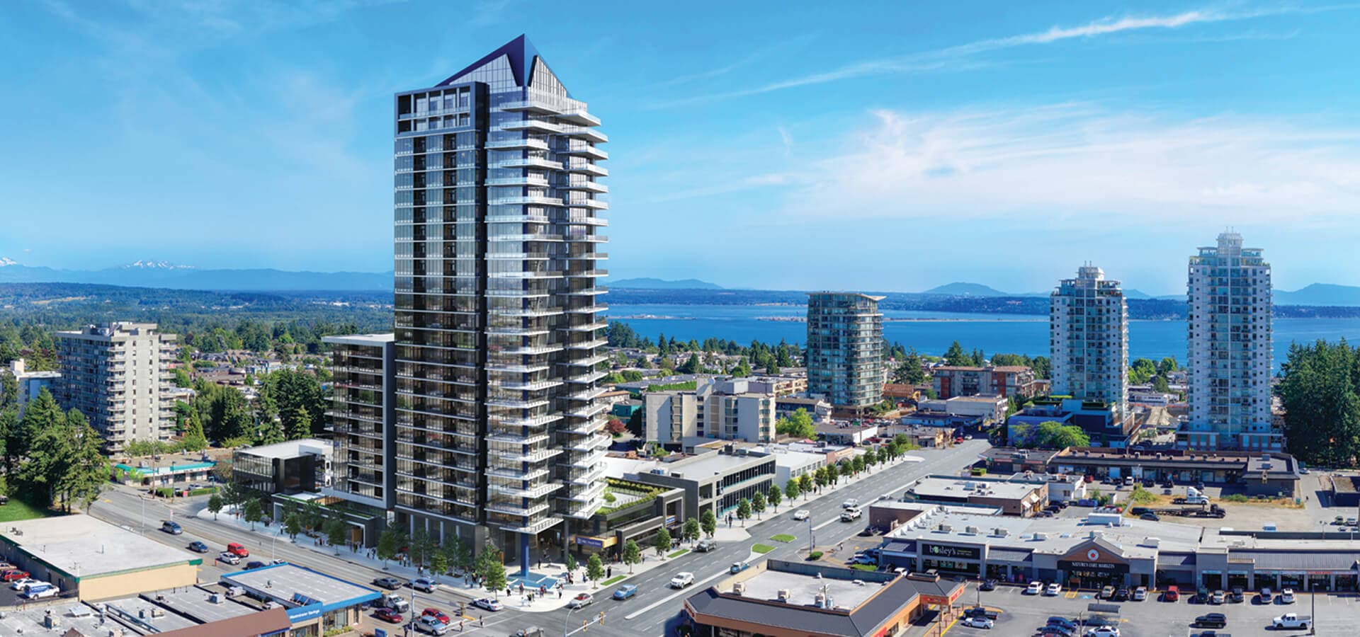 Guilden – An aerial daytime view of a tall glass high-rise tower with a distinctive angled crown, surrounded by a city street grid with shops, other towers, and water and mountains in the distance.