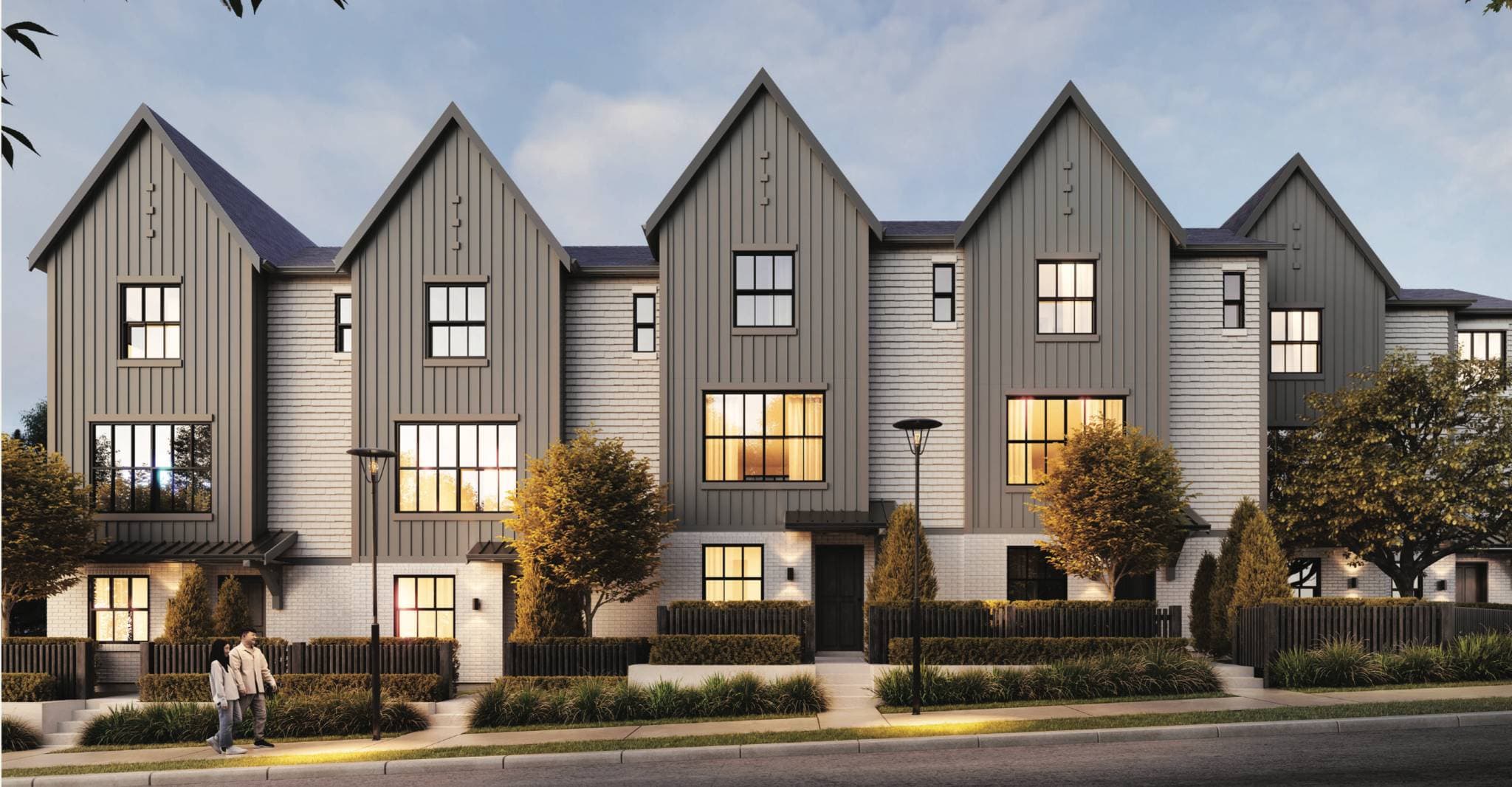 Renfrew South – Row of modern three-storey townhomes with steep gabled roofs, grey and white siding, and lit windows at dusk, with a couple walking on the sidewalk.