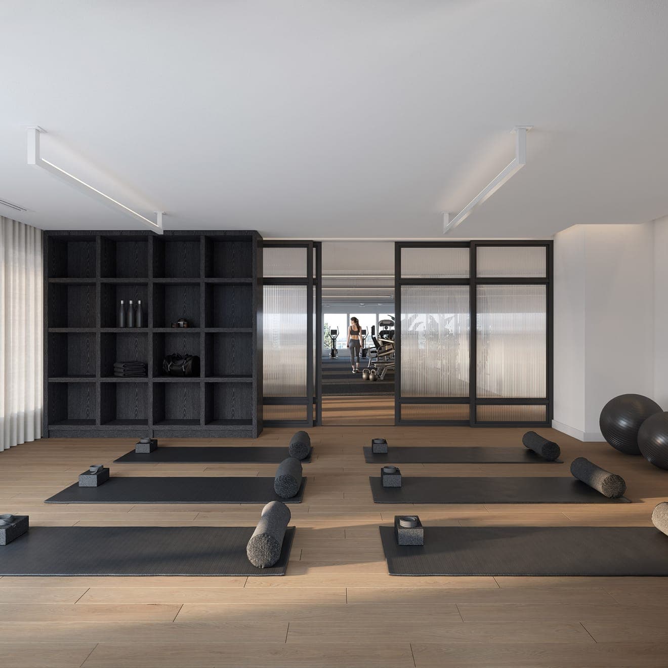 Smith & Farrow – A yoga studio with rolled mats and bolsters laid out on a wood floor, dark storage shelving, and a glass-walled gym visible beyond with a woman on a treadmill.