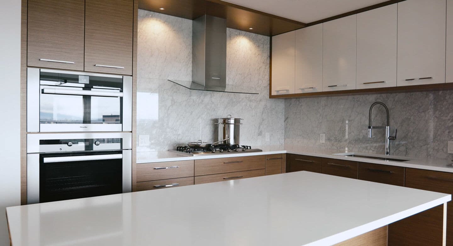 Smith & Farrow – A kitchen with wood and white cabinetry, marble backsplash, a gas cooktop, built-in double wall ovens, and an island in the foreground.