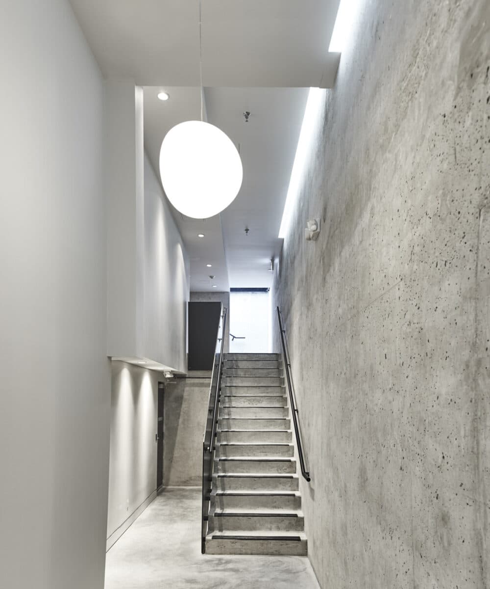 Naímo – Interior stairwell with concrete walls, a round pendant light fixture, and a staircase leading up to a lit doorway.