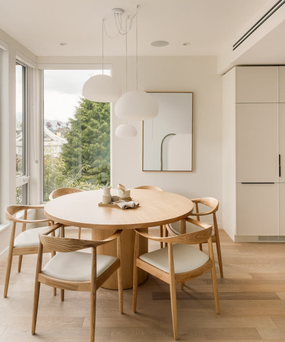 Naímo – Dining nook with a round wood table, matching wood chairs with cream cushions, hanging globe pendant lights, and a window view of trees.
