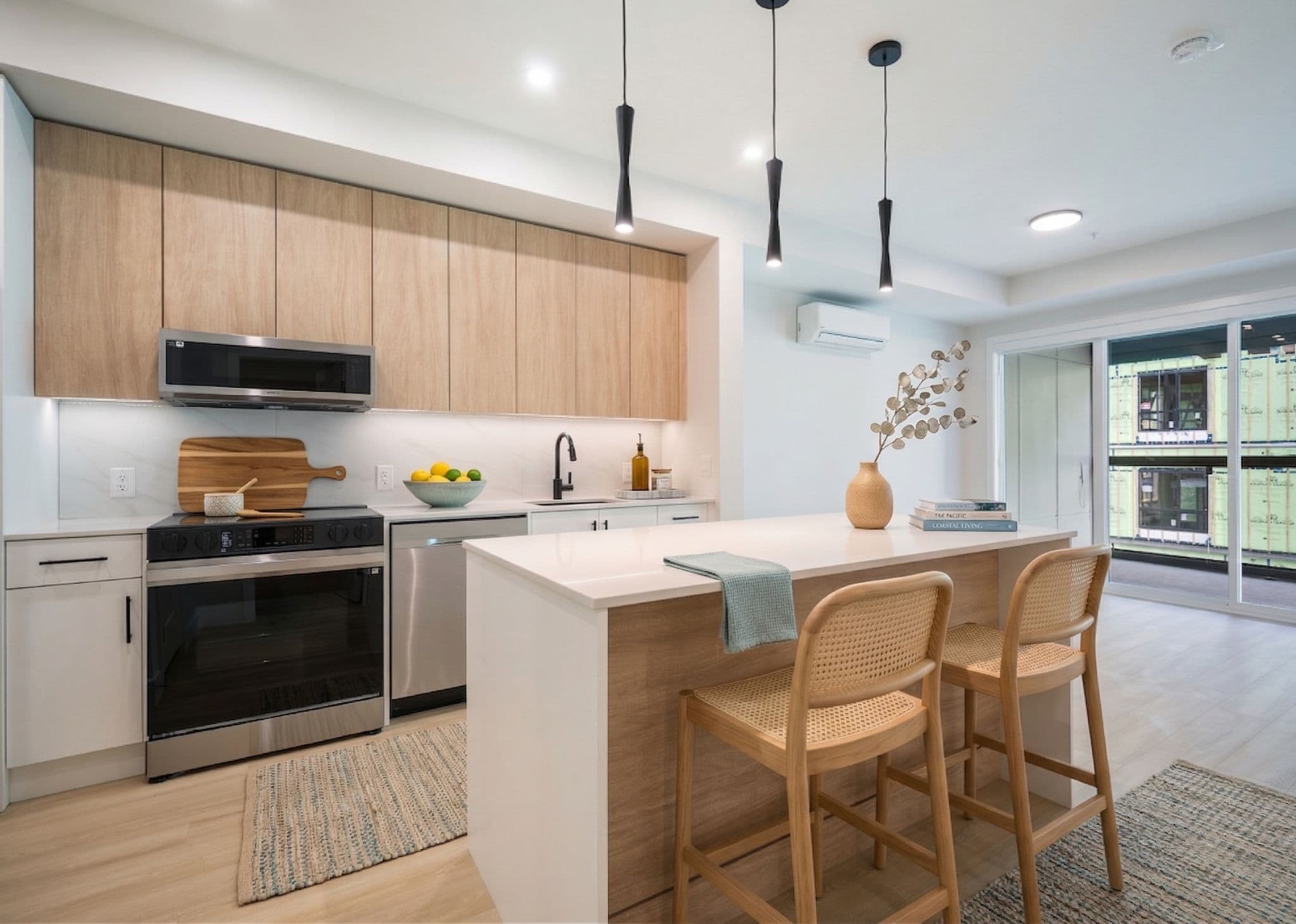 Sage 3 – Kitchen with light wood cabinetry, stainless microwave and stove, marble-look backsplash, and a view of an adjacent building under construction through the balcony door.