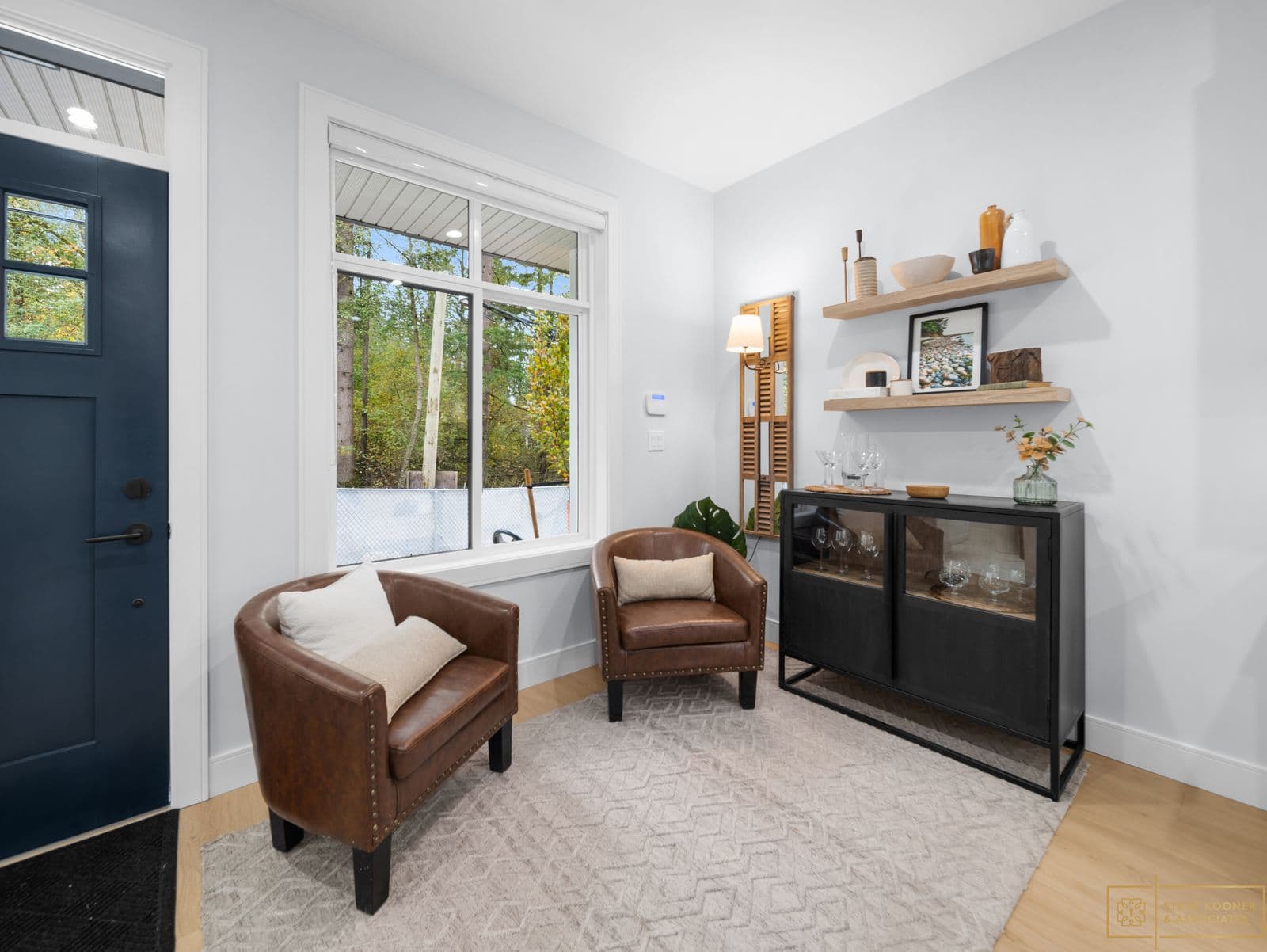 Raven's Park – Entryway sitting area with two brown leather accent chairs, a window looking onto a wooded yard, a black glass-front cabinet, and open wood shelving.