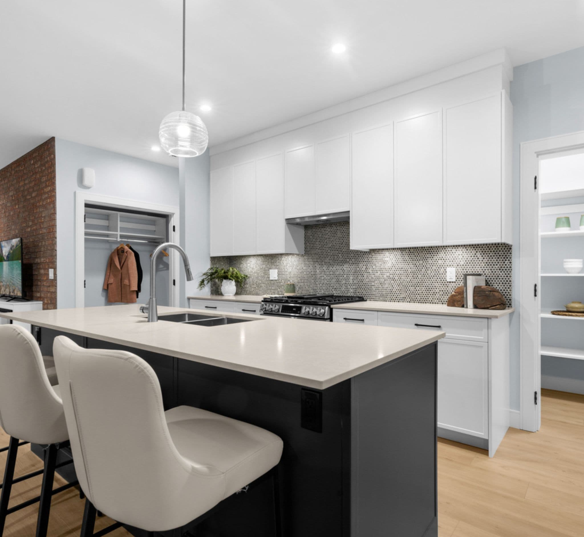 Raven's Park – Kitchen with white shaker cabinets, a penny-tile backsplash, stainless gas range, a dark island with bar stools, and a brick accent wall in the adjoining living area.