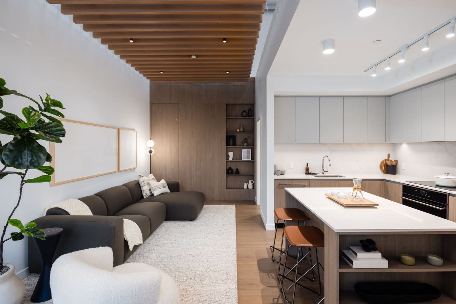 Koen – Living room with a wood slat ceiling, dark sectional sofa, and an open kitchen with gray cabinetry.