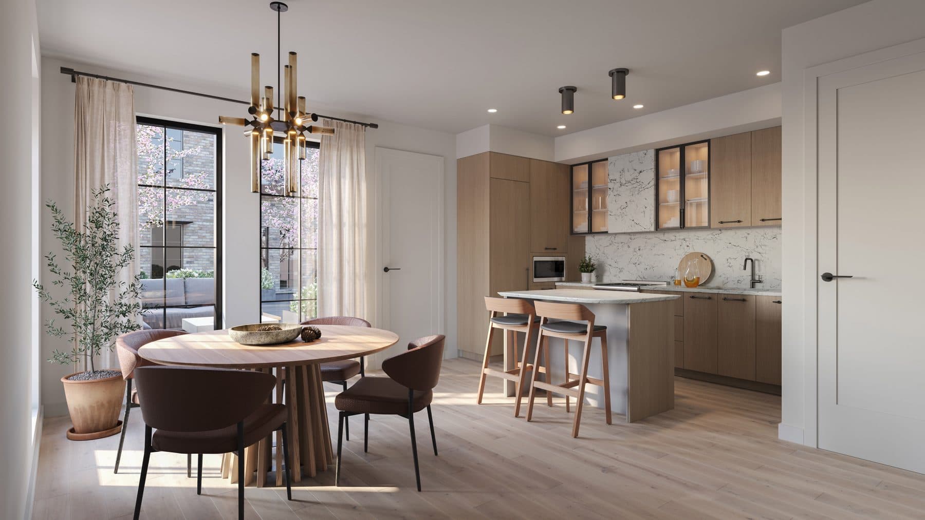 King and Columbia – An open-concept kitchen and dining area features light wood flooring, warm flat-panel cabinetry, a marble backsplash, and a modern island with two wooden bar stools. A round dining table with reddish-brown upholstered chairs sits under a brass chandelier next to large grid windows that reveal a sunny outdoor terrace with blooming trees.