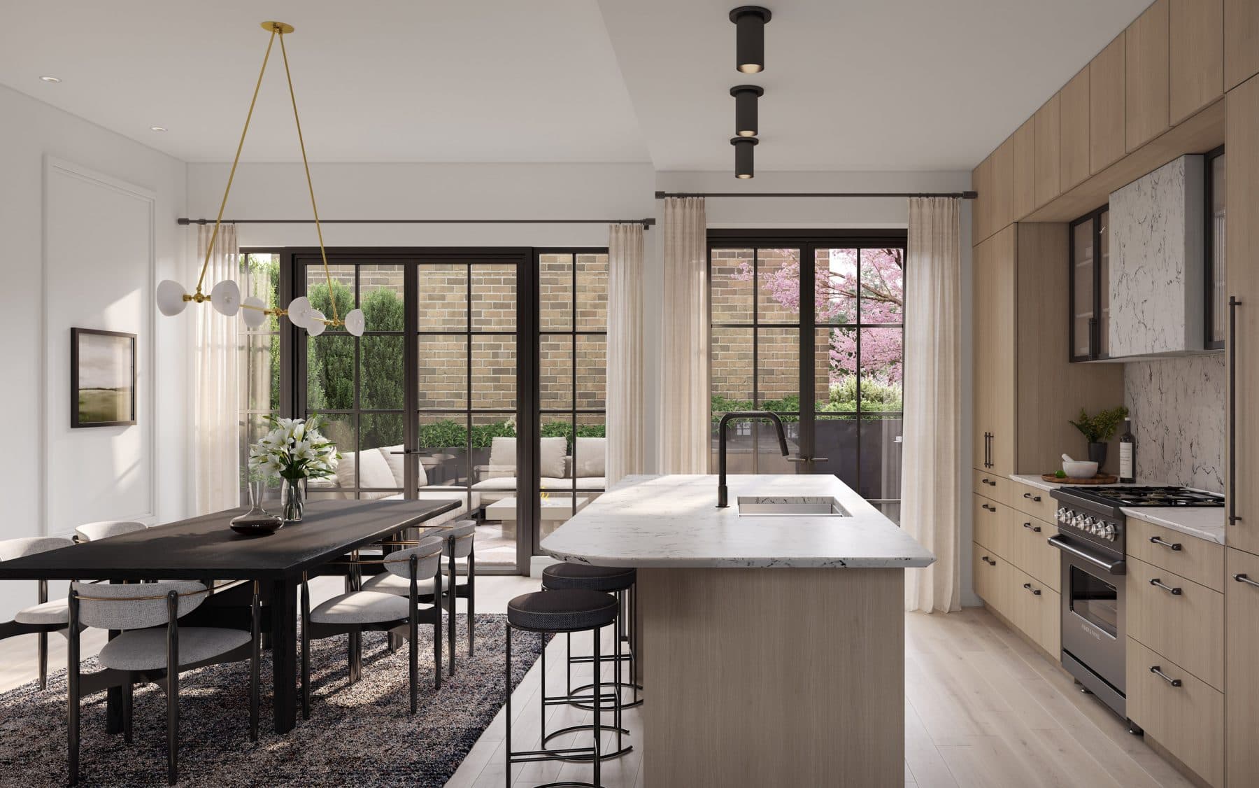 King and Columbia – An open-concept kitchen and dining space features a waterfall marble island with barstools, light wood cabinetry, and a dark wooden dining table beneath a modern brass chandelier. Large black grid-style sliding glass doors open out to a private brick courtyard patio with outdoor seating and a blossoming pink tree.