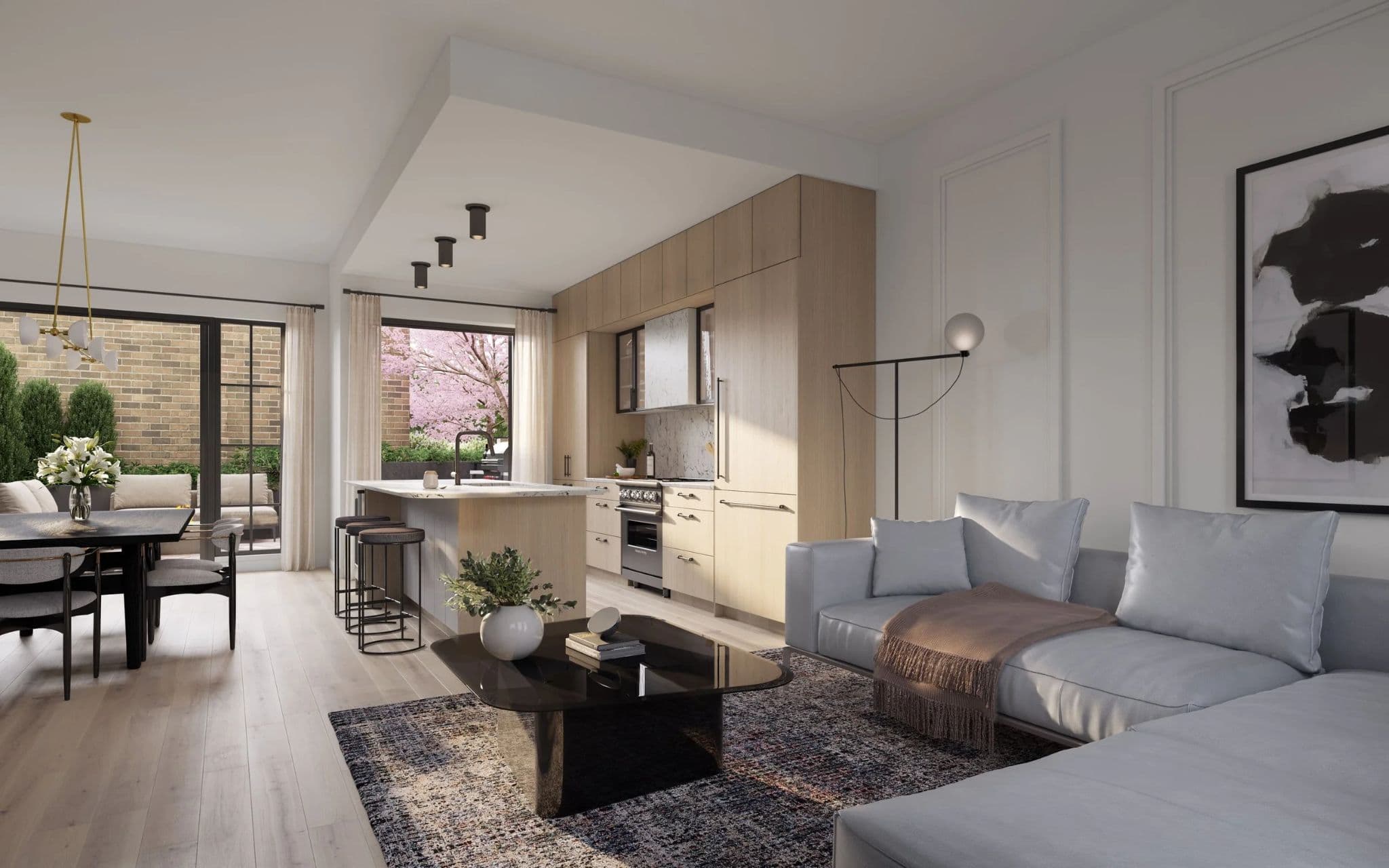King and Columbia – An open-concept living area and kitchen featuring light wood millwork, marble backsplashes, and light-toned hardwood flooring. Sunlight streams through glass doors that lead to a private patio, illuminating a light gray sectional sofa, dark coffee table, and an adjacent dining area.