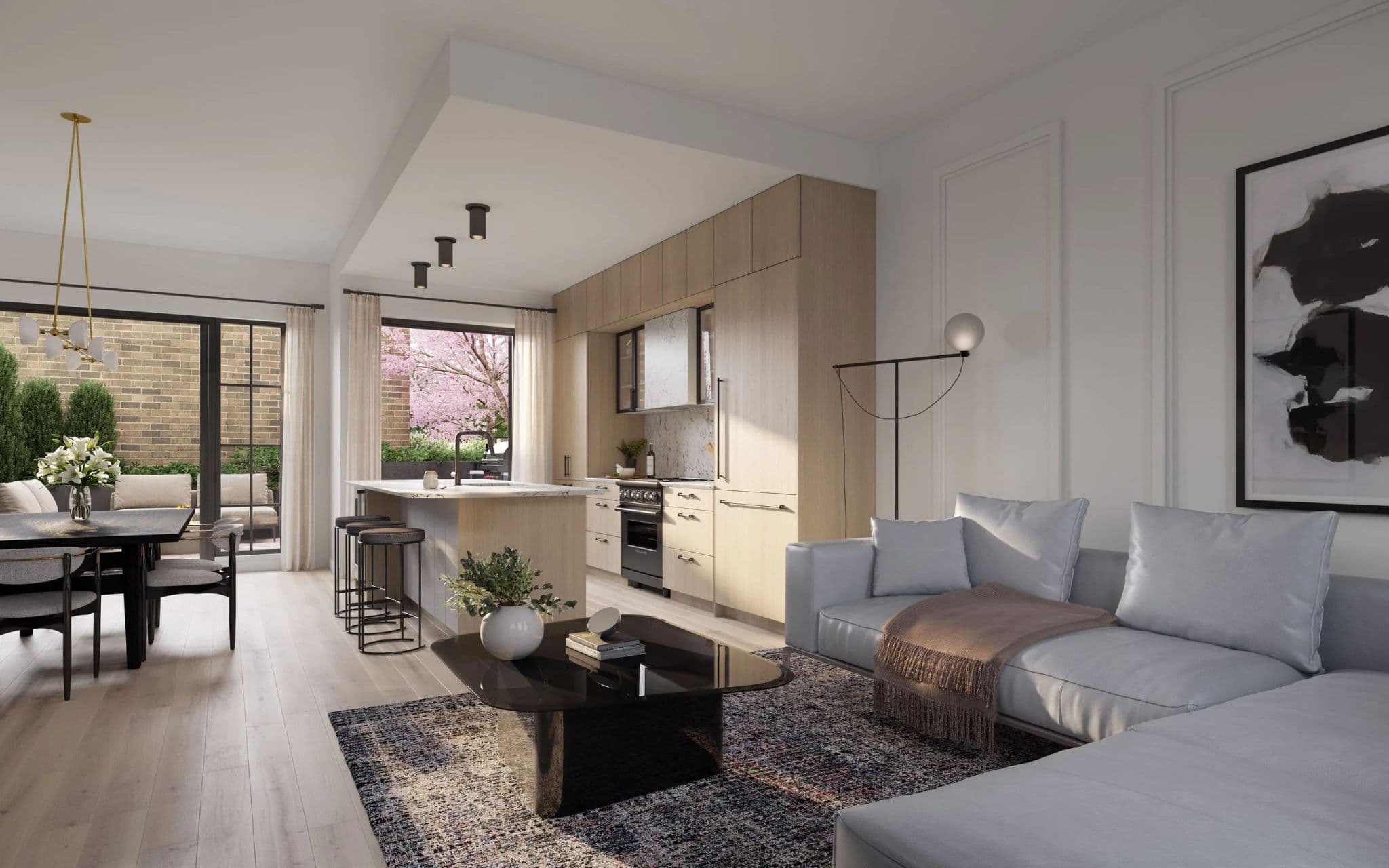 King and Columbia – A modern open-concept living area featuring a light-toned wood kitchen with a marble waterfall island and integrated appliances, alongside a bright living room anchored by a light blue sectional and abstract wall art. Glass patio doors at the back open onto a private outdoor garden space filled with lush greenery and a blooming pink tree.