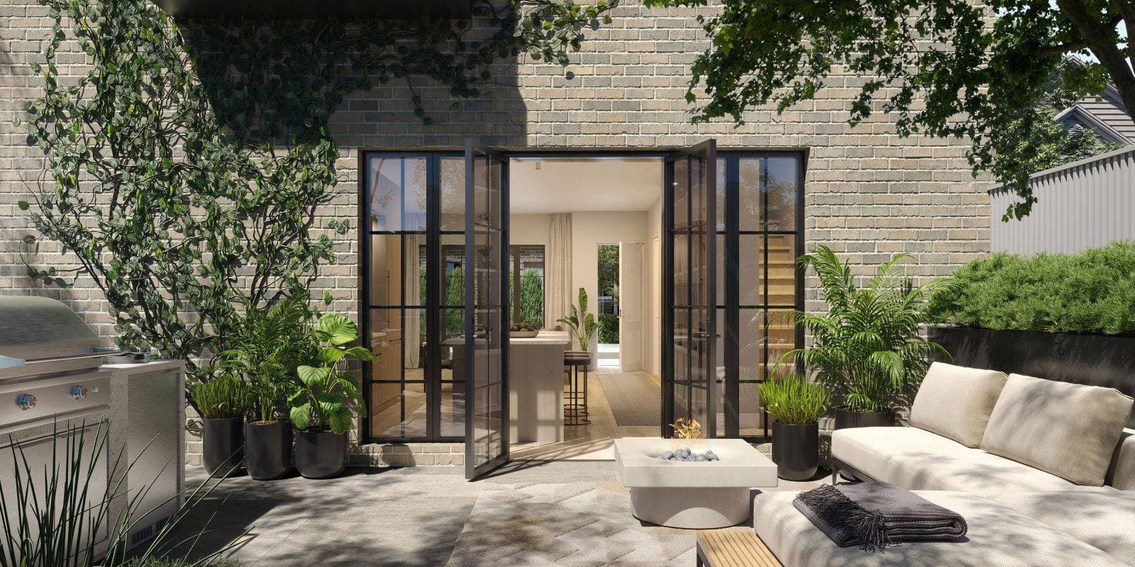 King and Columbia – A sunny private patio featuring a light grey brick facade, climbing green ivy, and black-framed French doors open to the interior kitchen and living space. The outdoor space is furnished with a built-in stainless steel BBQ grill, potted tropical plants, a modern low-profile fire pit, and a cushioned lounge daybed under tree shade.