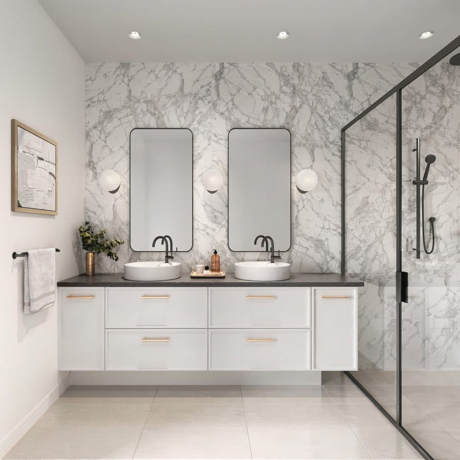 King and Columbia – A modern bathroom features a floating white vanity with gold hardware, dual vessel sinks, matte black gooseneck faucets, and twin pill-shaped mirrors flanked by globe sconces. The accent wall and glass-enclosed walk-in shower are wrapped in dramatic grey-veined white marble tiles under soft recessed lighting.