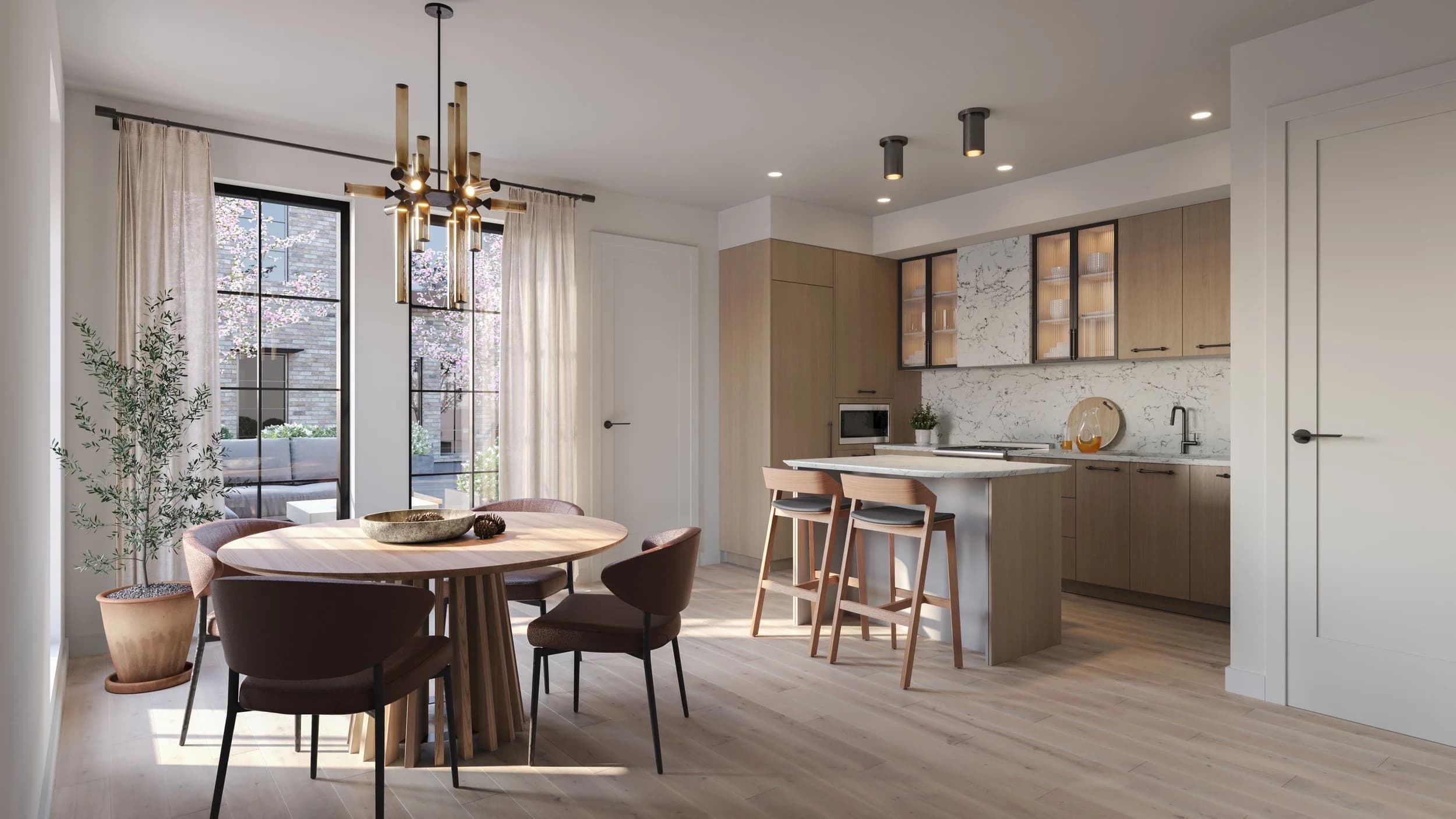 King and Columbia – An open-concept kitchen and dining area featuring light wood flooring, flat-panel wood cabinetry, marble backsplash, and a central kitchen island with two barstools. Natural light streams in through large floor-to-ceiling windows next to a round wooden dining table, revealing a view of a courtyard with blooming pink trees outside.