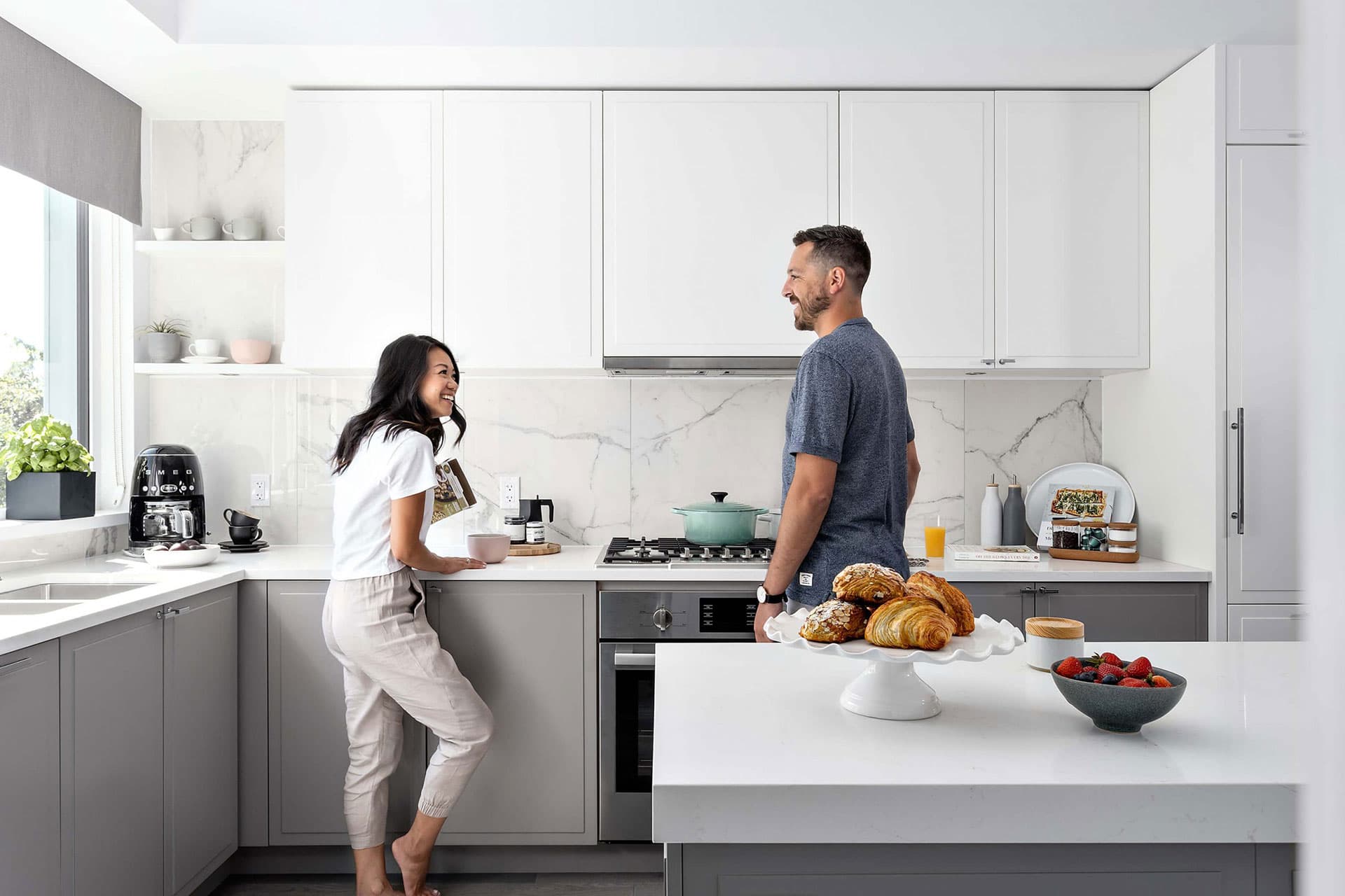 York – A modern white-and-grey kitchen with marble backsplash where a man and woman stand talking near a waterfall-edge island with pastries and fruit.