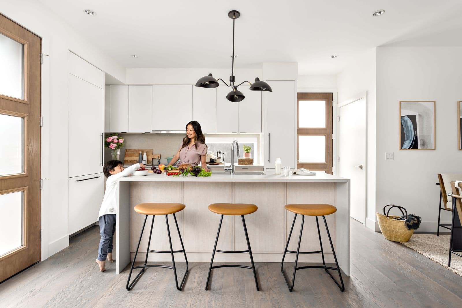 York – A bright white kitchen with a large island, pendant lighting, and leather-topped bar stools; a woman and child prepare food together.
