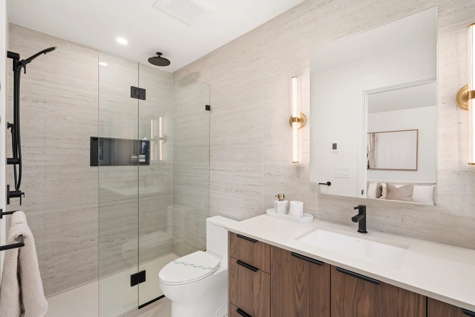 WE Killarney – A modern bathroom interior featuring light beige textured wall tiles, a glass-enclosed walk-in shower with matte black fixtures, and a toilet. A warm wood vanity with a white countertop is paired with a matte black faucet, flanked by sleek brass wall sconces alongside a large frameless mirror.