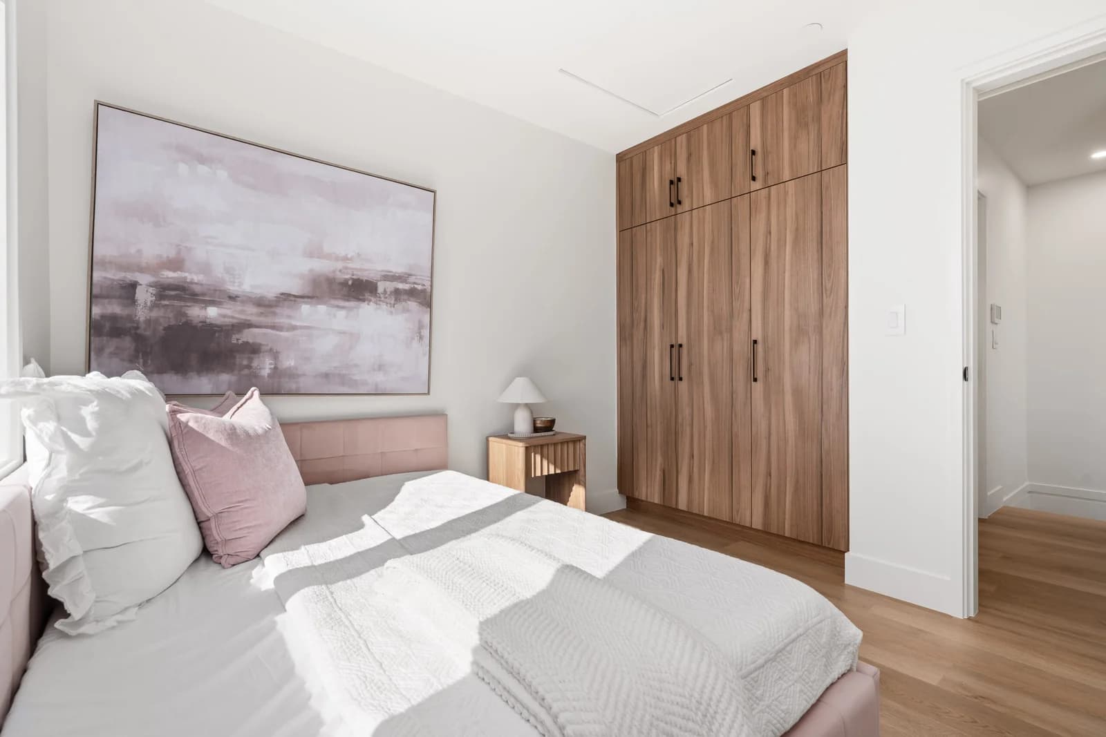 WE Killarney – A sunlit, modern bedroom features a bed dressed in white linens and blush pink accents alongside a small wooden nightstand. A wall of floor-to-ceiling built-in wood grain closets provides storage against crisp white walls and light wood flooring.