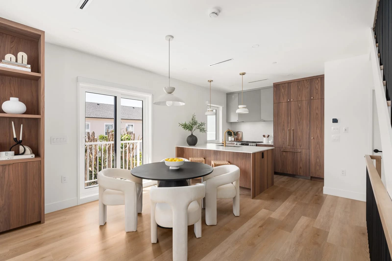 WE Killarney – A modern open-concept dining area and kitchen featuring light hardwood floors, crisp white walls, and rich walnut cabinetry. A round black dining table surrounded by curved white chairs sits in the foreground, adjacent to sliding glass doors that open to a balcony, with a contemporary kitchen island and built-in walnut cabinetry behind it.