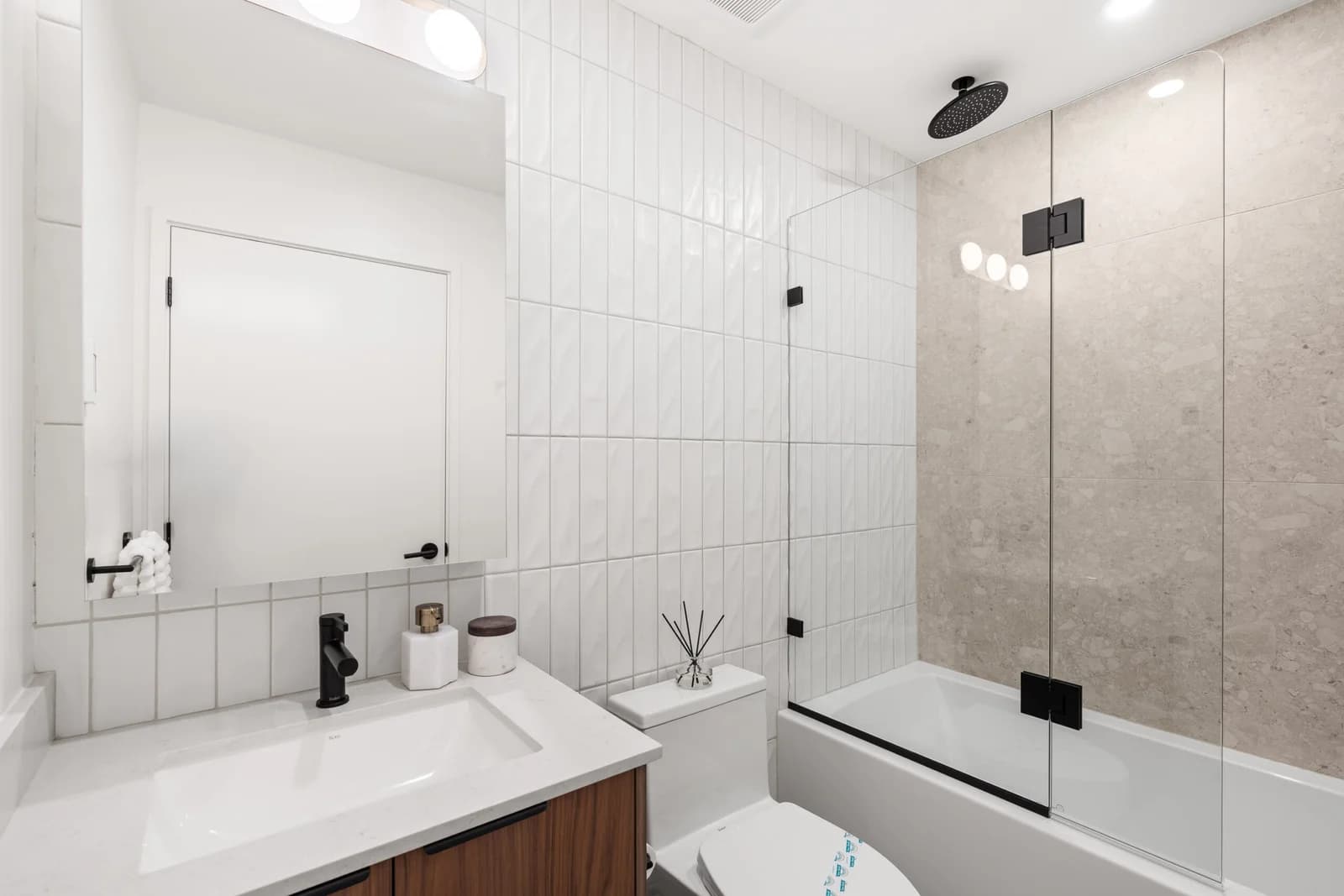 WE Killarney – A modern, brightly lit bathroom featuring a white vanity with matte black fixtures, an undermount sink, and a large mirrored medicine cabinet. Adjacent is a white toilet and a combination bathtub-shower enclosed by a sleek glass partition, set against white vertical wall tiles and neutral stone-look tiles.