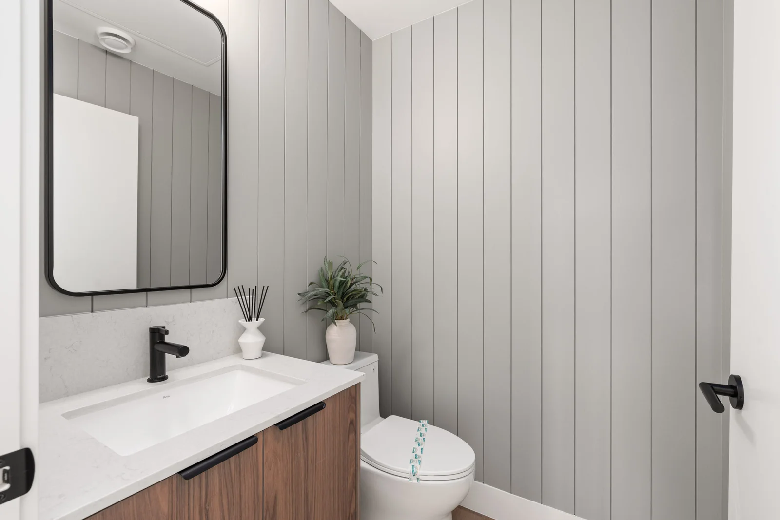 WE Killarney – A modern powder room featuring light grey vertical shiplap walls, a white toilet, and a warm wood vanity with a white countertop. Black hardware accents include a sleek matte black faucet, cabinet pulls, and a rounded rectangular mirror above the sink.