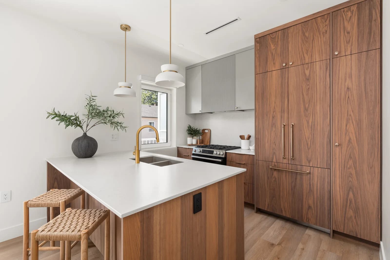 WE Killarney – A modern kitchen featuring rich walnut wood cabinetry, integrated appliances, and a central island with white quartz countertops and a brushed gold gooseneck faucet. Two white pendant lights hang above the island alongside woven counter stools, and daylight streams in from a window overlooking a nearby building.