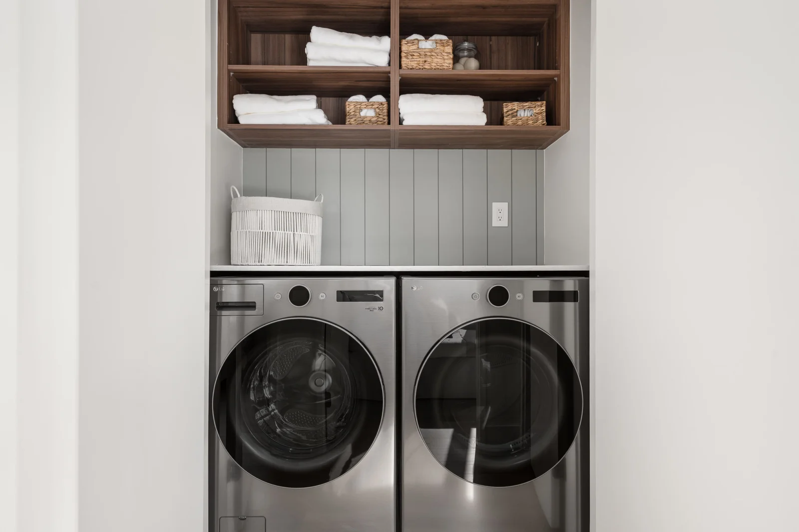 WE Killarney – A clean, modern laundry nook features a side-by-side stainless steel washer and dryer set fitted neatly beneath a countertop. Above, light grey vertical shiplap backs open dark wood shelving neatly staged with folded white towels and woven storage baskets.