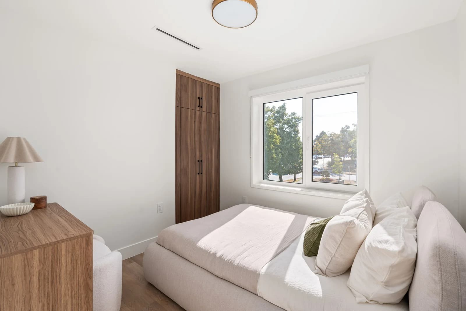 WE Killarney – A bright, minimalist bedroom features a low-profile upholstered bed dressed in neutral linens beside a large window that lets in abundant daytime sunlight. A tall, built-in dark wood closet and matching wooden dresser contrast cleanly with crisp white walls and light wood flooring.