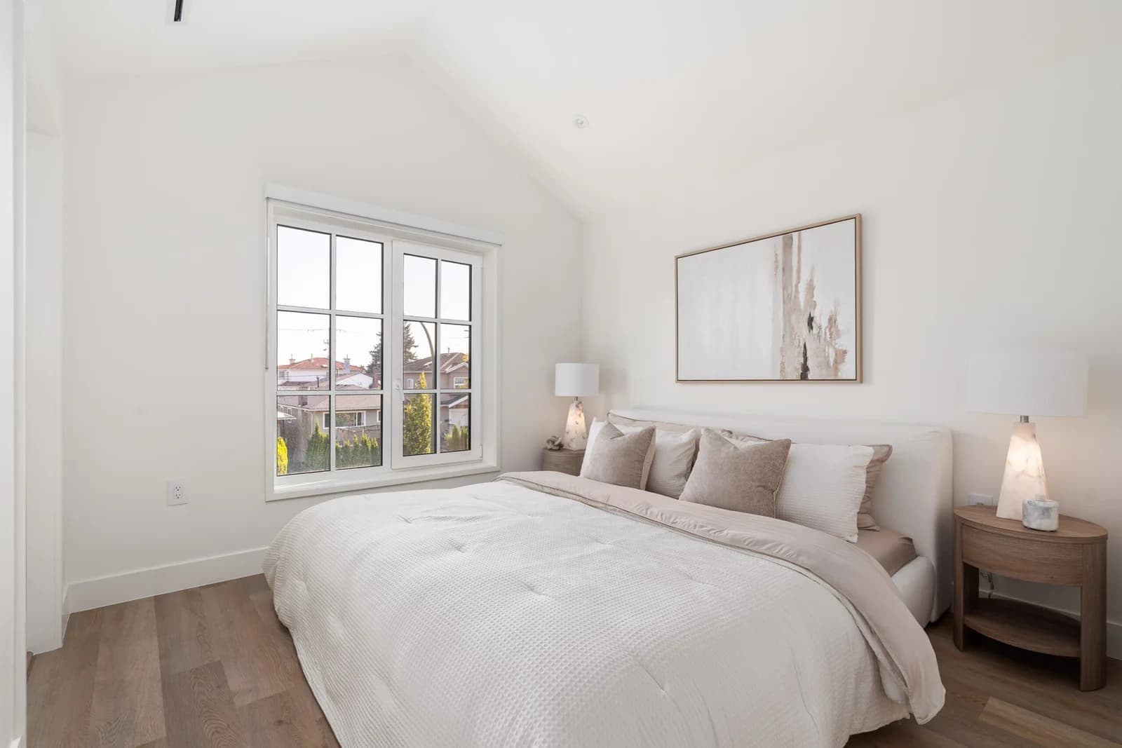 WE Killarney – A bright, minimalist bedroom features a vaulted white ceiling, light hardwood floors, and a neatly made bed with neutral textured linens. Flanking wooden nightstands hold modern table lamps, while a large multi-pane window lets in ample daylight and overlooks a residential neighborhood.