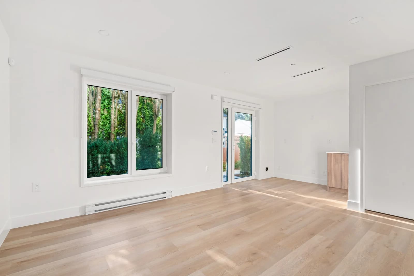 WE Killarney – An unfurnished, sunlit room features light-toned hardwood floors, crisp white walls, and a large window looking out onto lush green foliage. Glass patio doors lead outside, with natural sunlight casting bright geometric patterns across the wooden planks.