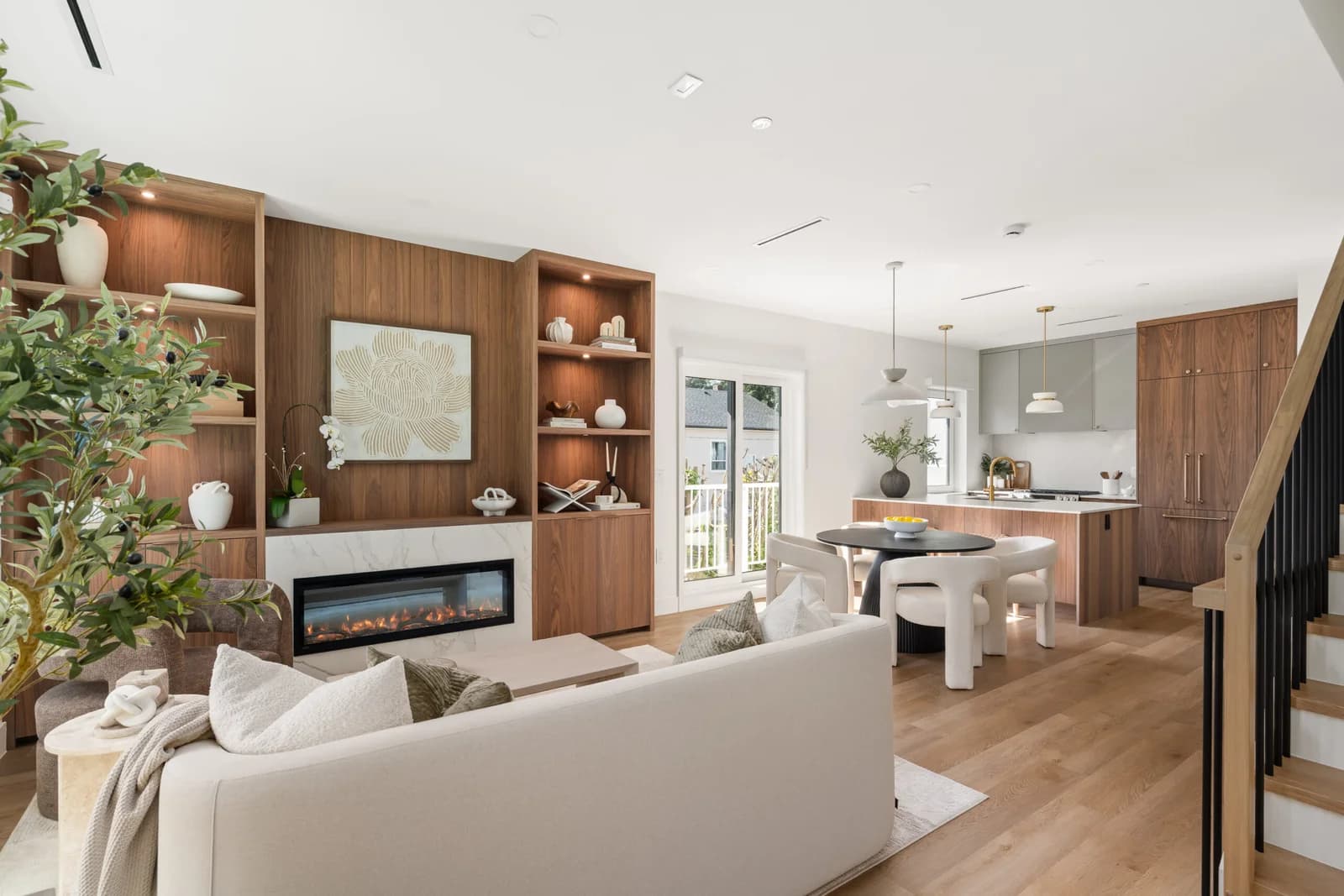 WE Killarney – A bright, open-concept living, dining, and kitchen area featuring light wood floors and custom warm wood built-ins framing a linear fireplace with marble surround. Natural light streams in through glass patio doors beside a modern round dining table and a fully equipped kitchen with wood and grey cabinetry.
