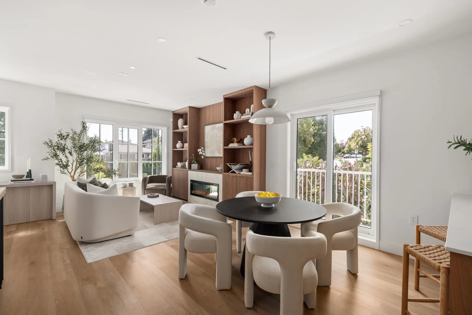 WE Killarney – A bright, open-concept living and dining area featuring light hardwood floors, white walls, and a round black dining table surrounded by sculptural cream upholstered chairs. In the background, a cozy living room includes a white curved sofa, custom wood built-in shelving with an integrated fireplace, and large windows and glass sliding doors letting in ample natural daylight.