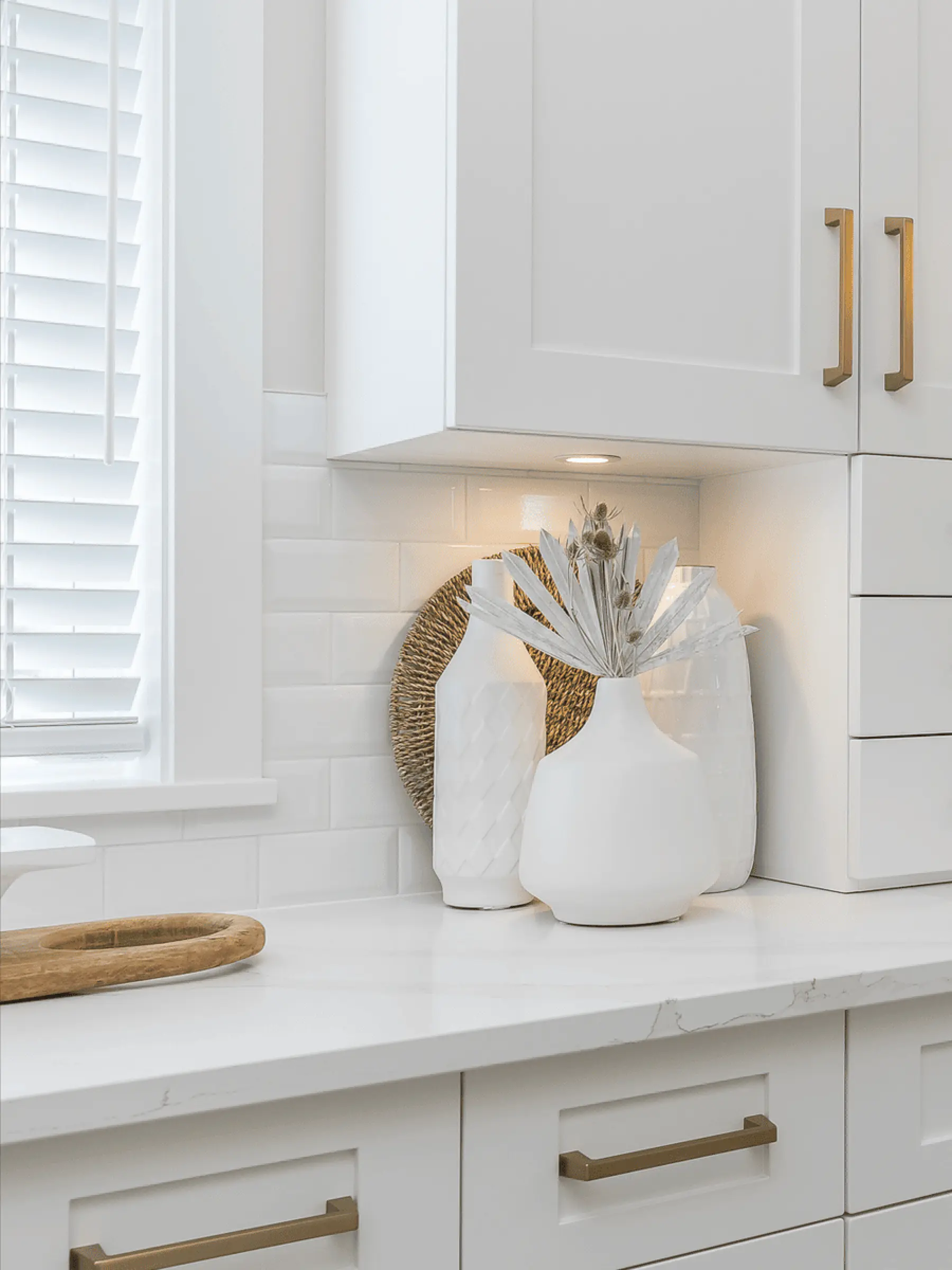 Malloway Village – A bright kitchen corner featuring white shaker-style cabinetry with brass handles, white subway tile backsplash, and light-veined quartz countertops. Decorative white ceramic vases with dried botanicals sit under warm under-cabinet lighting next to a window with white horizontal blinds.