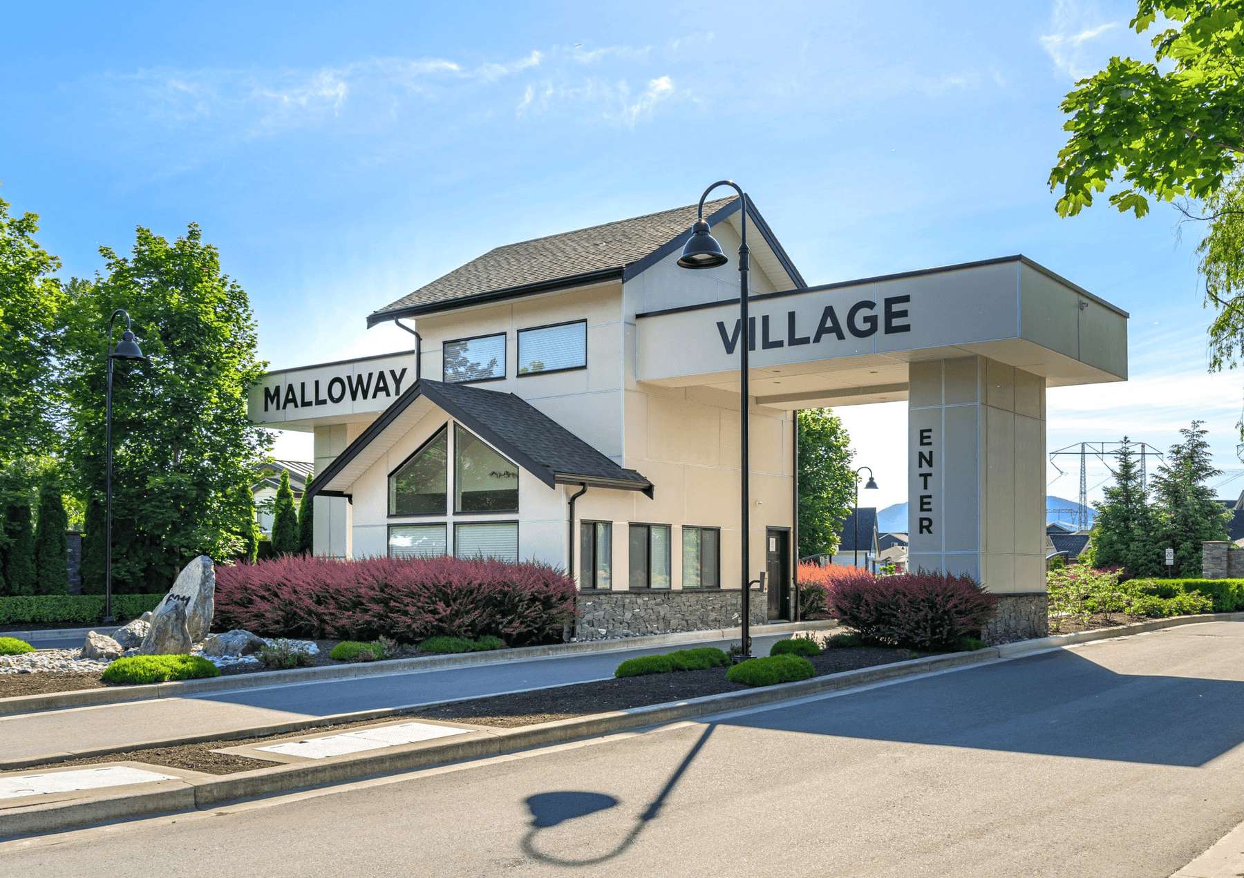 Malloway Village – A gated community entrance featuring an overhead gatehouse structure with white siding, stone base trim, and dark-trimmed pitched roofs alongside the road. Manicured bushes, landscaped rock features, and vintage-style lampposts flank the driveway under a bright blue sky.