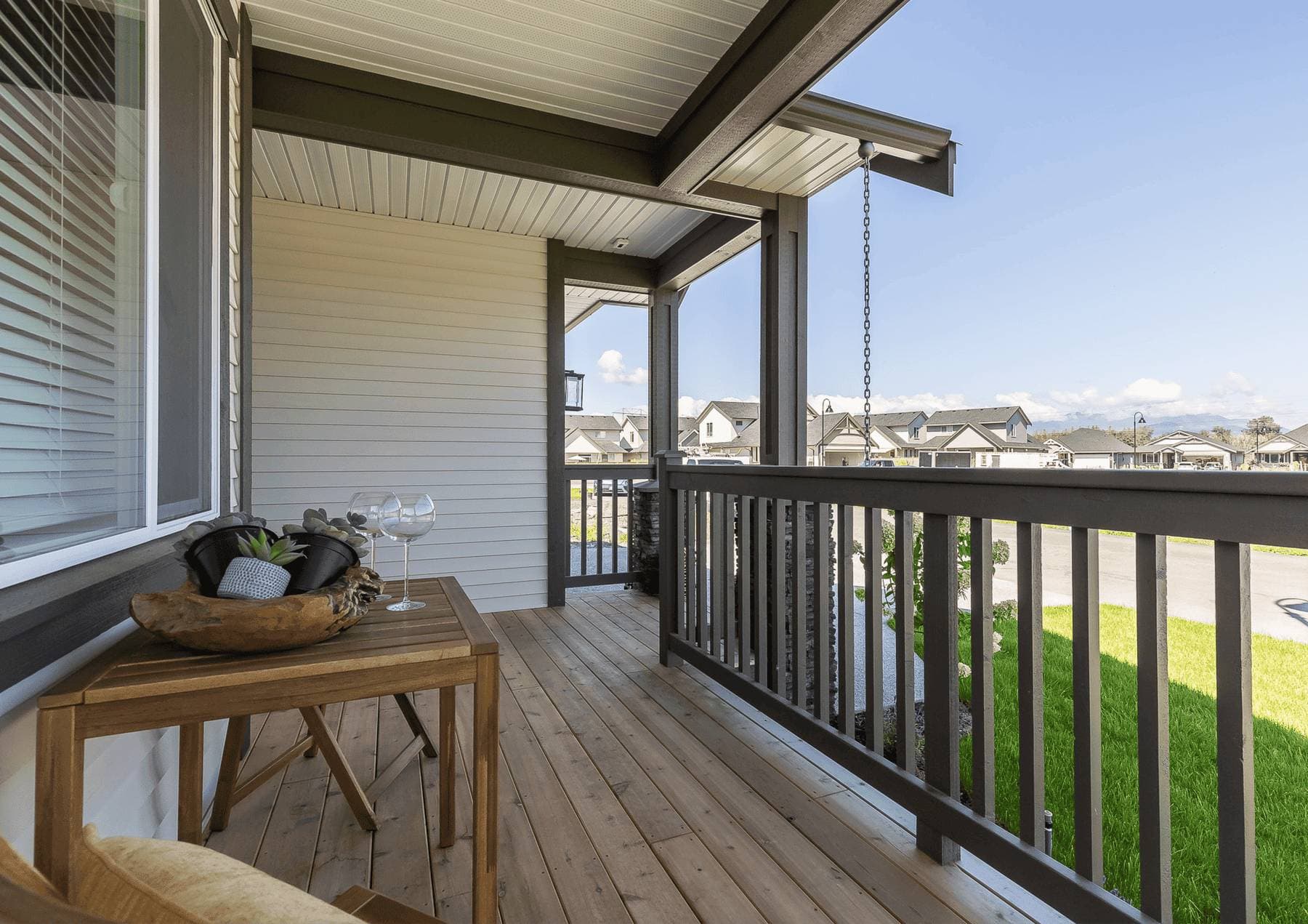 Malloway Village – A covered front porch features light wood plank decking, dark grey railings, and a small wooden side table styled with wine glasses and a potted succulent. Beyond the porch railing, the view looks out onto a tidy suburban street with green lawns and matching contemporary single-family homes under a clear blue sky.